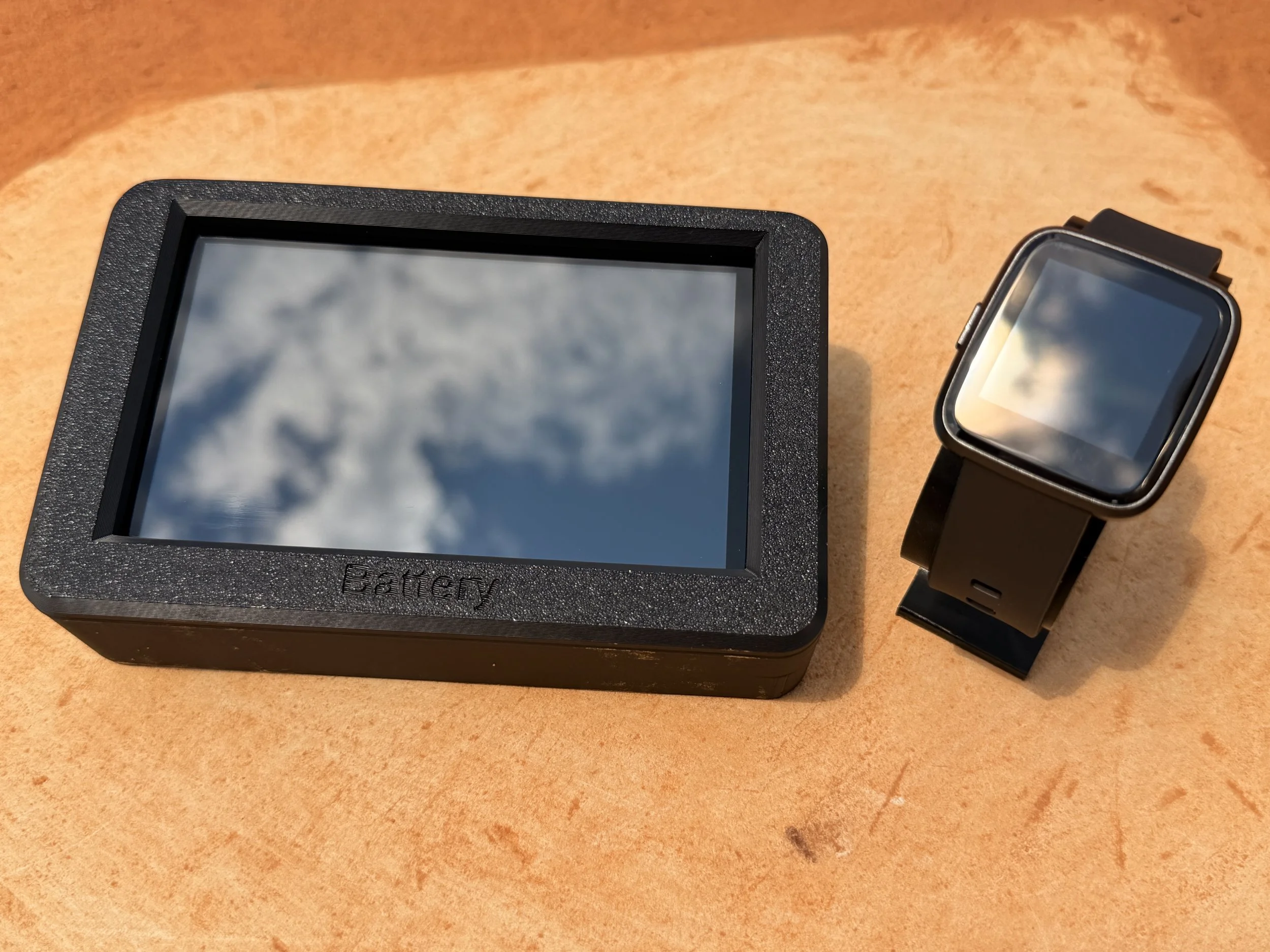 A black electronic device with a screen next to a smartwatch on a tan surface.