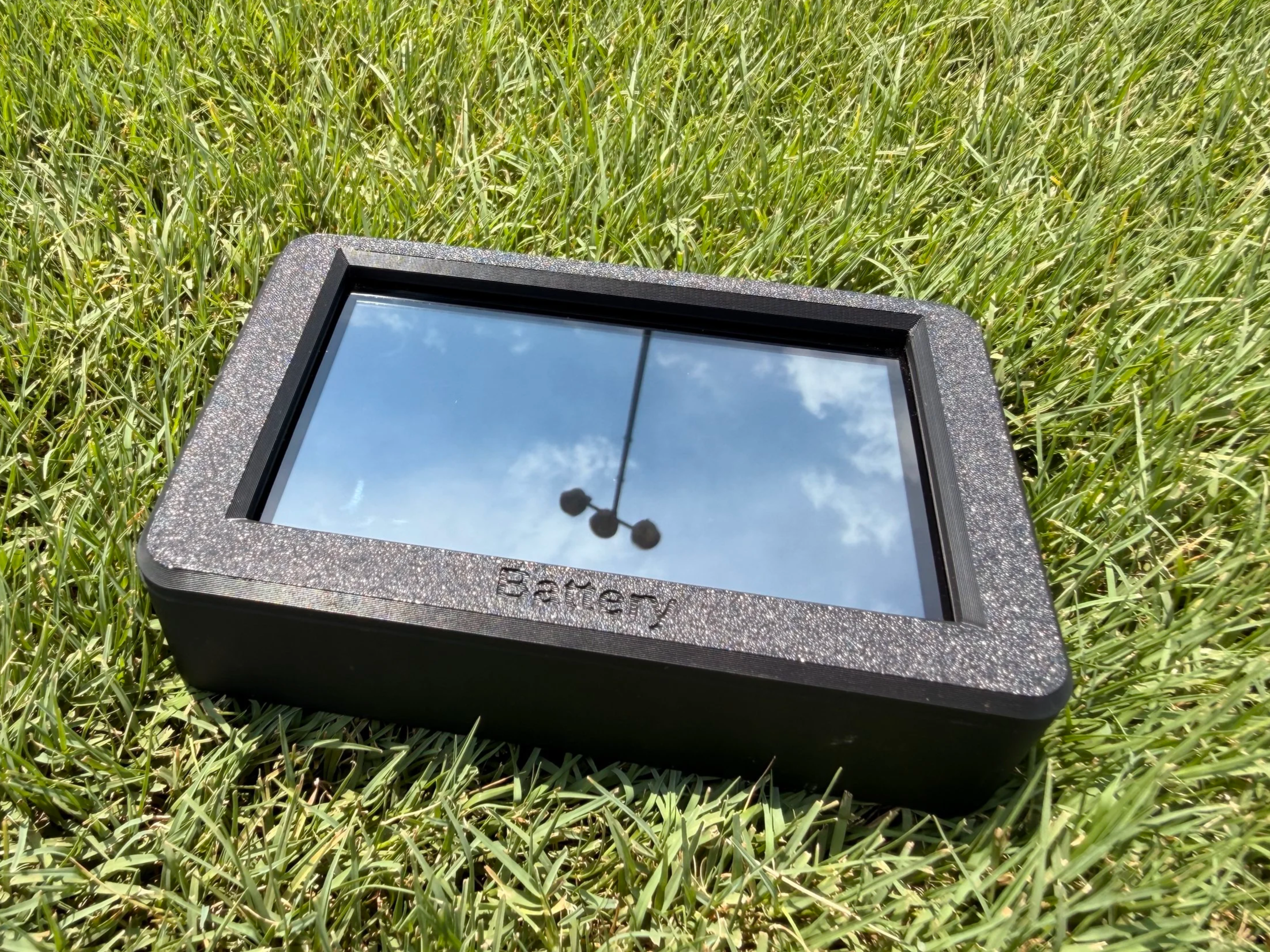 A small, black, rectangular reflective device with a glittery or sparkly border placed on green grass, reflecting the sky and a street lamp.
