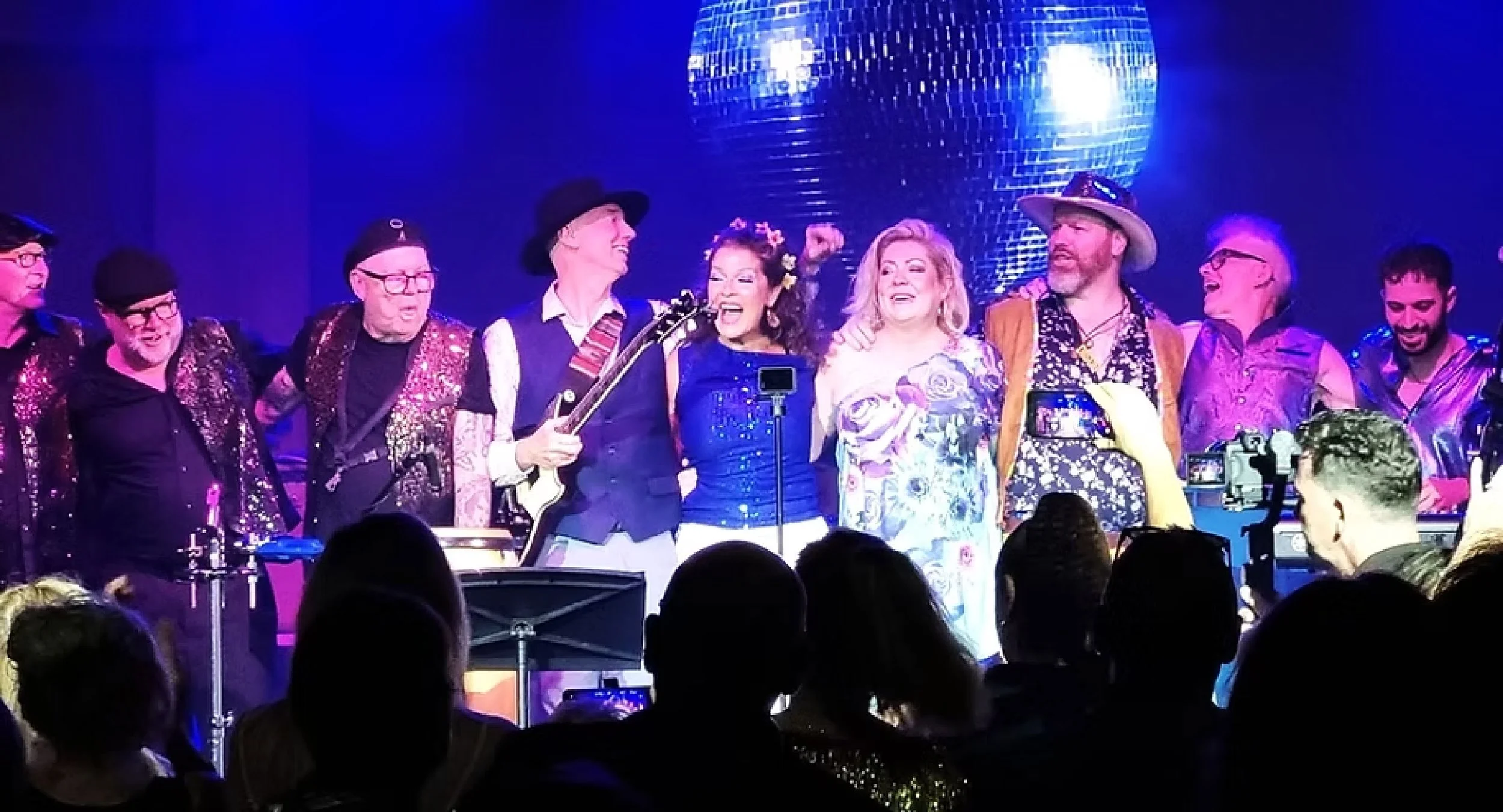 A group of performers on stage with a large disco ball overhead, smiling and celebrating, with audience members in the foreground taking photos.