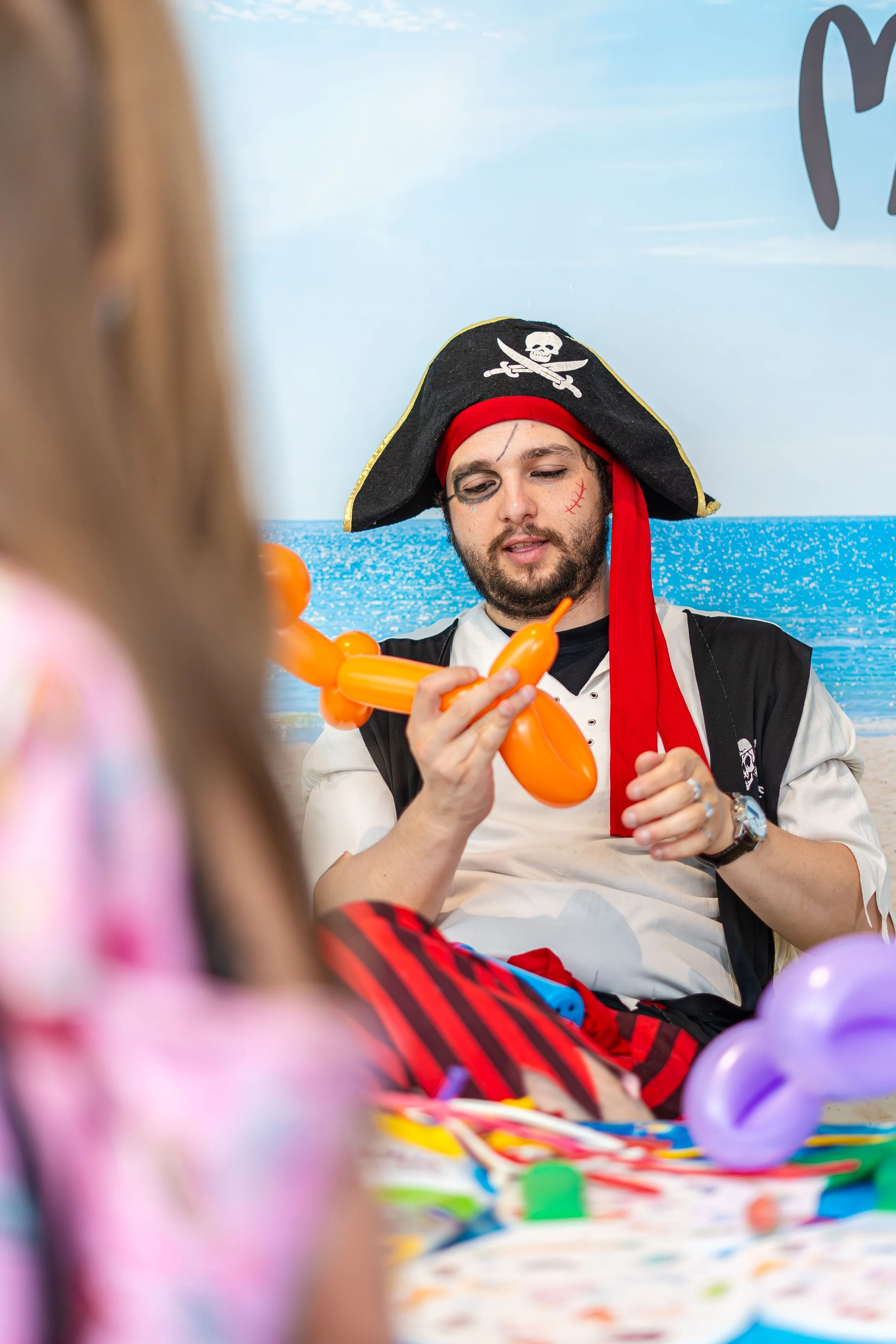Person dressed as a pirate with a painted face, wearing a pirate hat with a skull and crossbones, and a red bandana, sitting at a table with colorful balloons and decorations, engaging with a child, with the ocean and sky in the background.