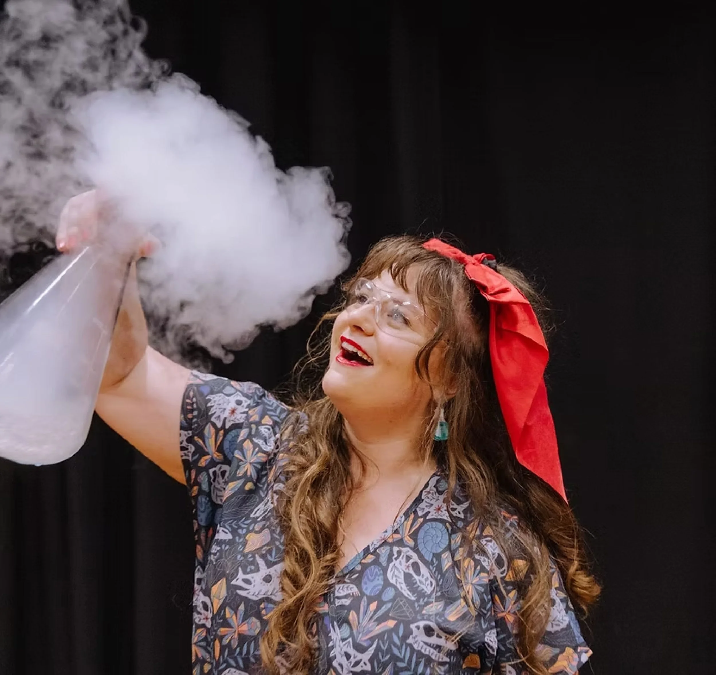 Woman with long curly hair wearing glasses and a colorful patterned dress, smiling and holding a clear container filled with dry ice, with white fog emitting from it, against a black background.