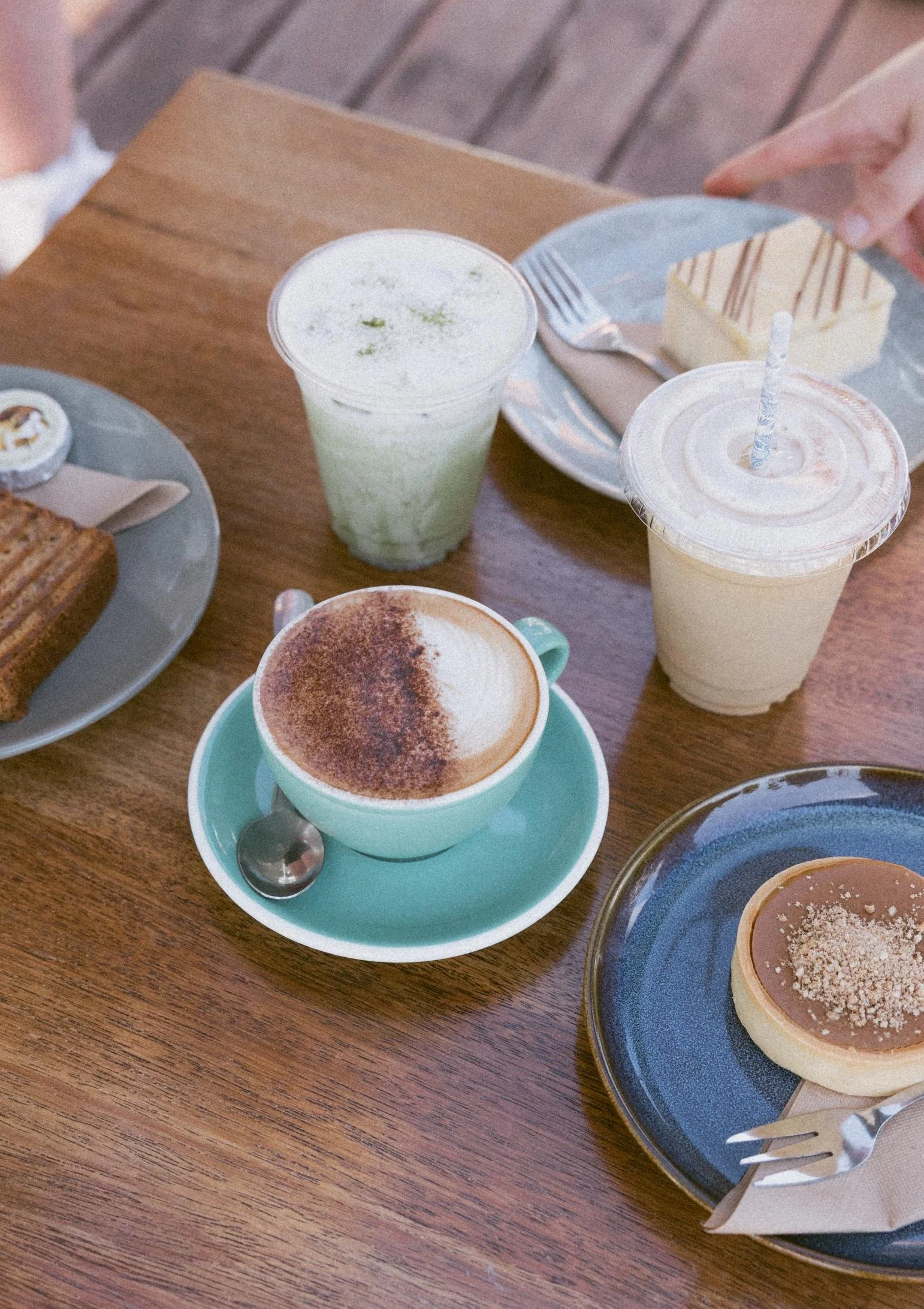Various drinks and desserts on a wooden table, including matcha, an iced coffee, cappuccino and a caramel tart.