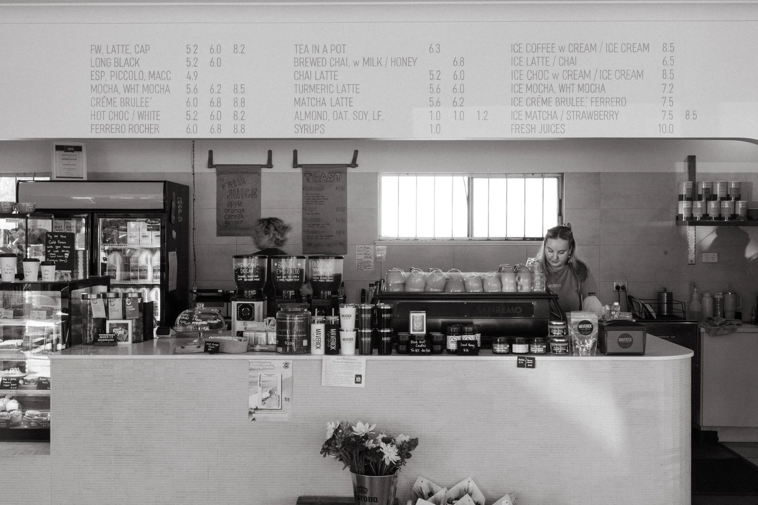 The image shows the interior of a coffee shop with a menu board above the counter listing various coffee and tea options, prices, and syrups. There are two employees behind the counter, and various coffee-making equipment and supplies are visible. In the foreground, there is a potted flower and some informational posters.