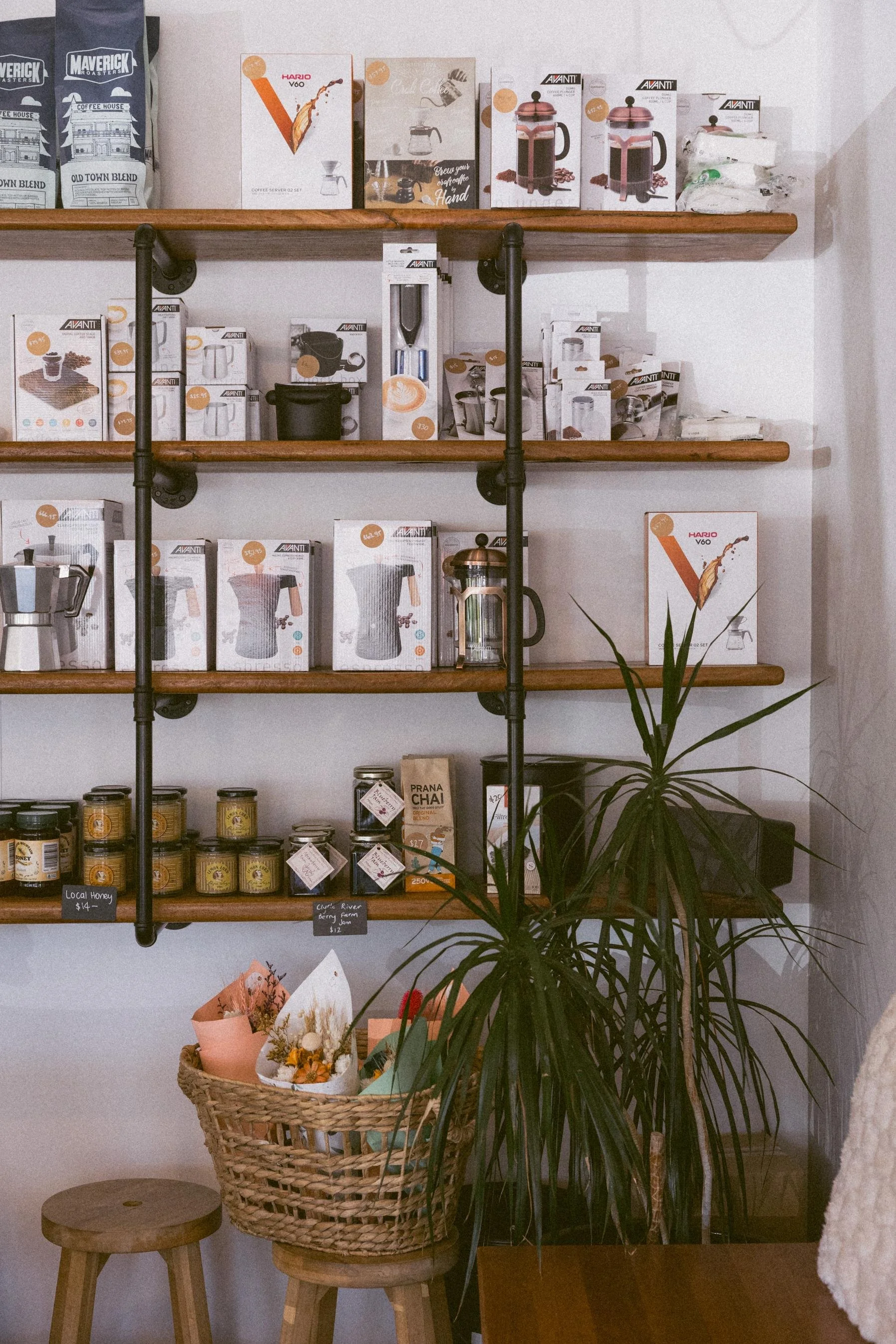 Interior of a store with wooden shelves displaying coffee and tea products, some labeled from local farms, and a plant in a basket on stools in front.