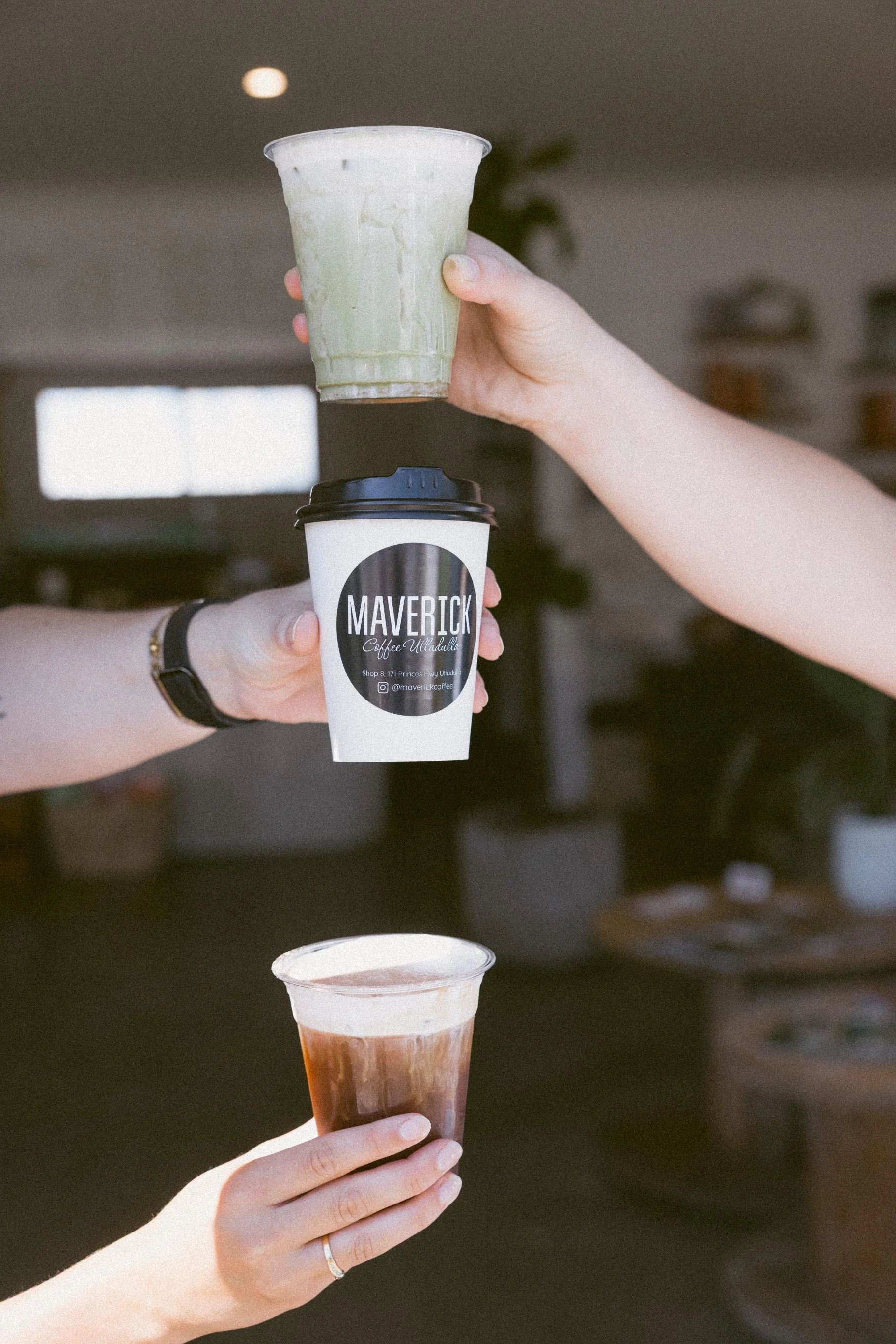 Three people are holding various drinks: one with a green iced beverage (Matcha), one with a hot coffee cup labelled 'MAVERICK', and one with an iced coffee in a plastic cup. The background is indoors with windows and some blurred furniture and plant