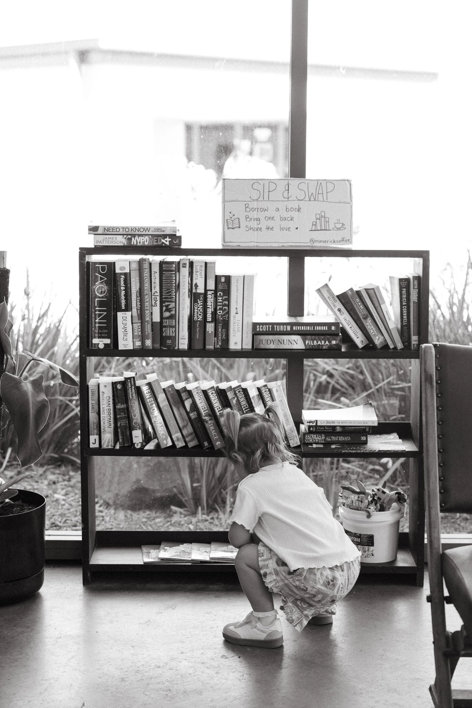 Child crouching and looking down, in front of a bookshelf filled with books.. There is a sign above the bookshelf for a book exchange program and a window with a bright outdoor scene in the background.