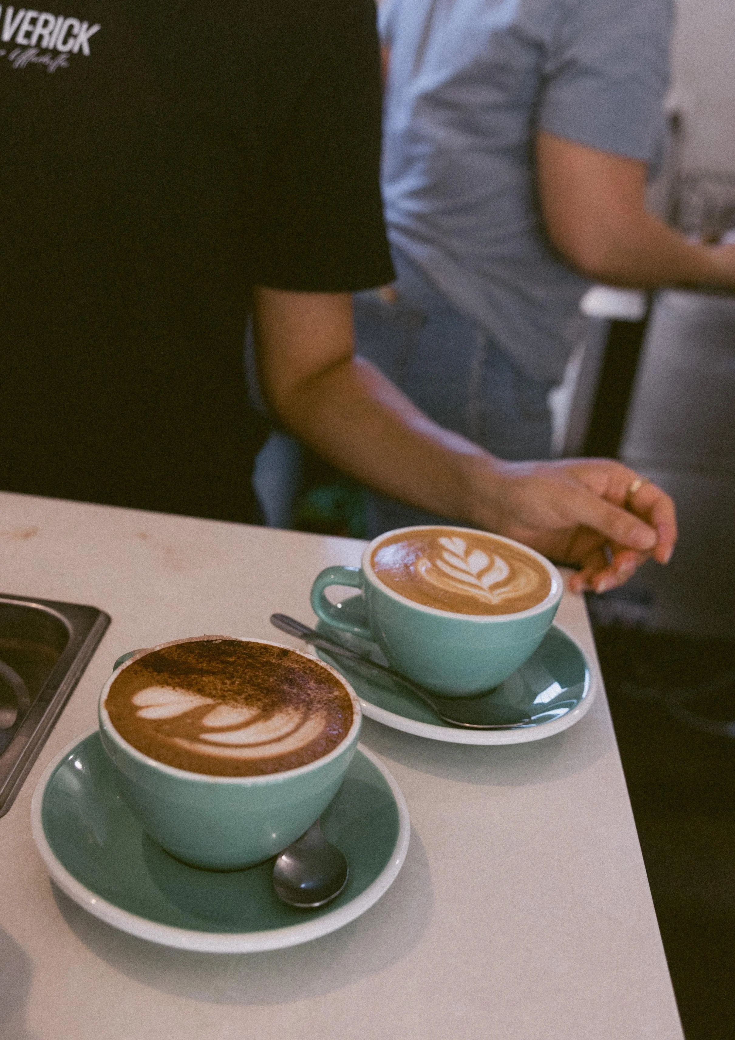 Two cups of coffee with latte art on a white table, with people sitting nearby. One person is wearing a black shirt, and another person is wearing a light blue shirt.