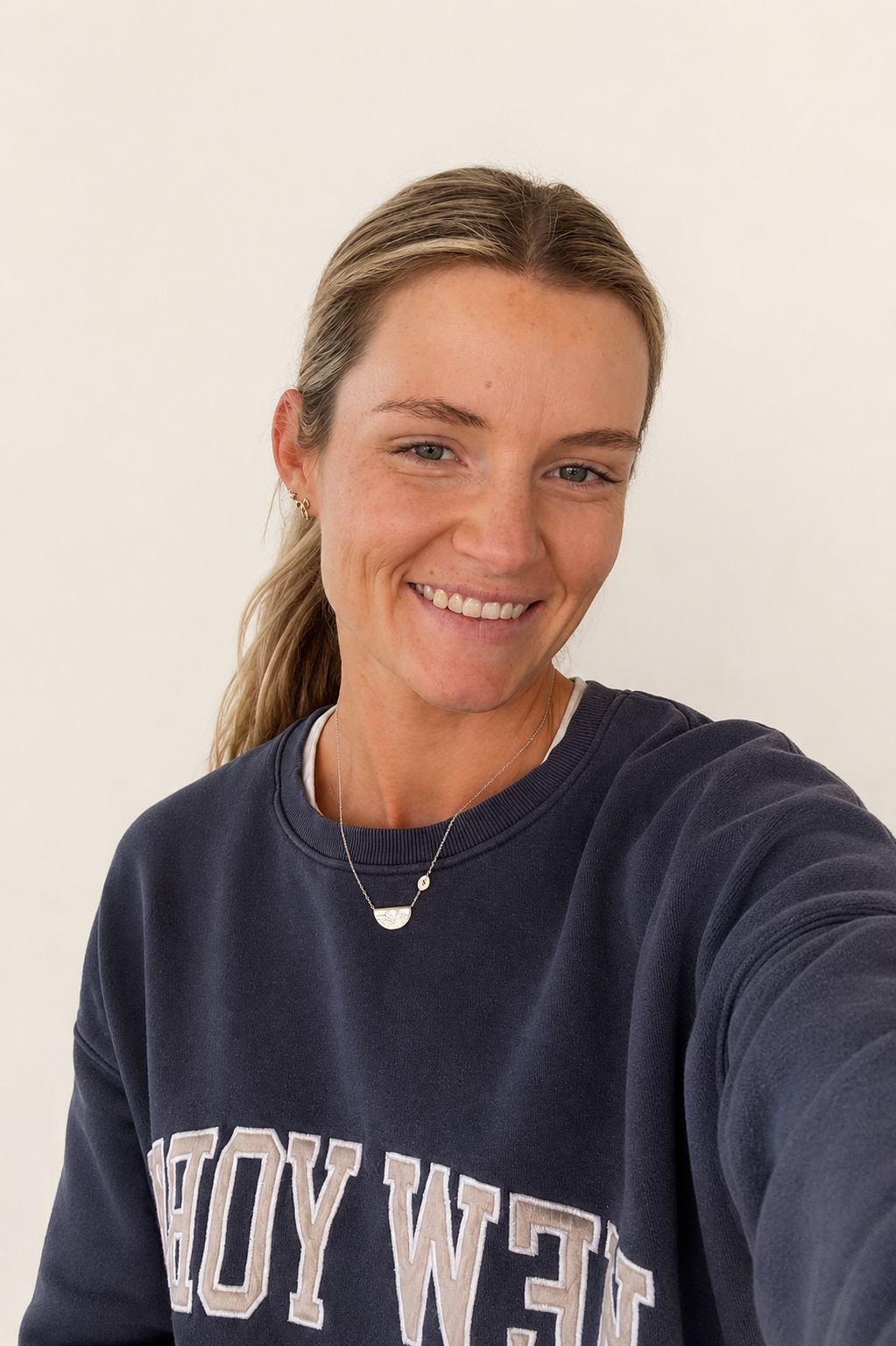 A young woman with blonde hair in a ponytail smiling and taking a selfie against a plain white background, wearing a navy blue sweatshirt with white text and layered necklaces.