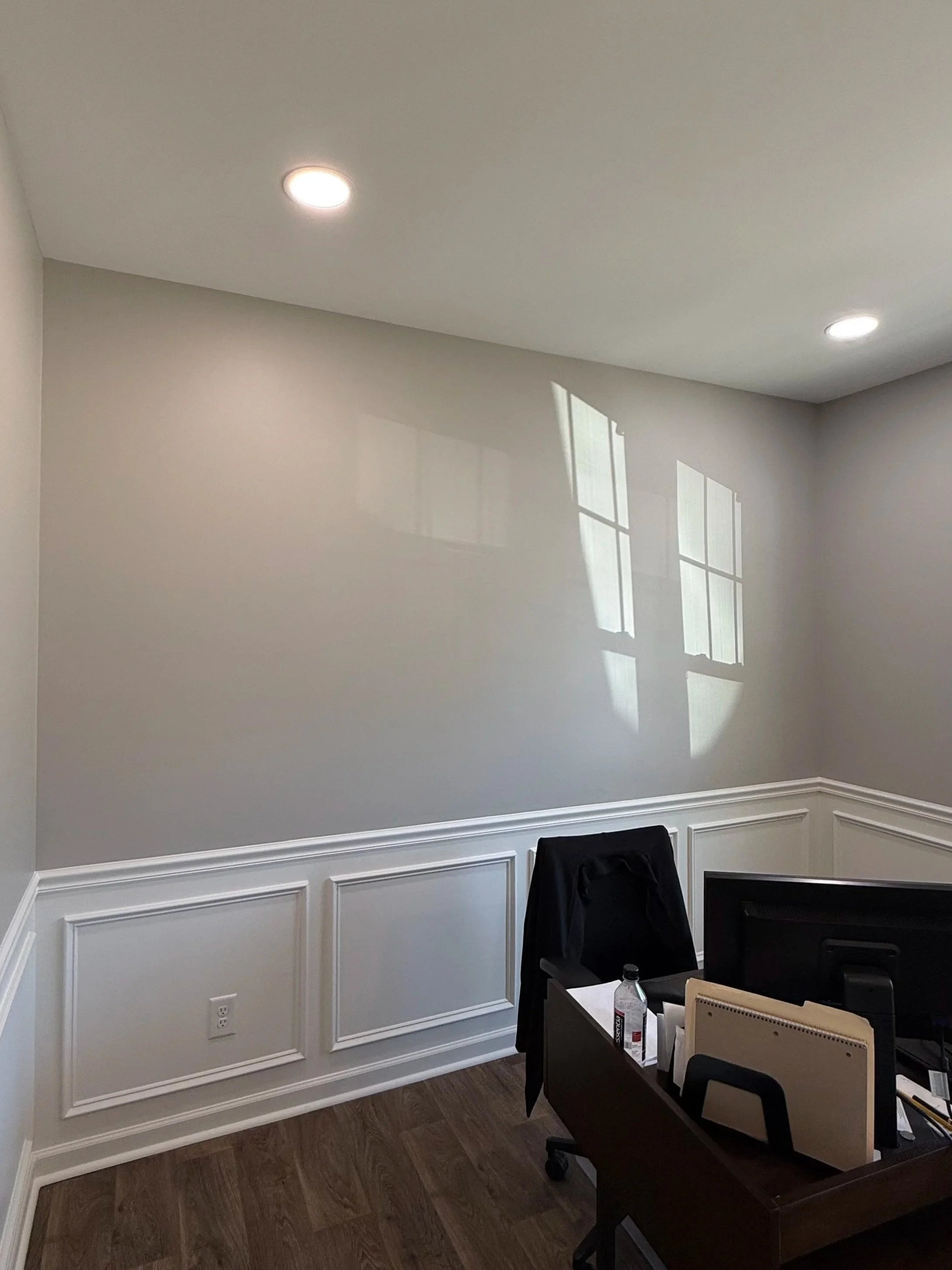 Office room with gray walls, white wainscoting, hardwood floors, two ceiling lights, a desk with a computer monitor, a water bottle, and a chair with clothing draped over it. A window casts a shadow on the wall.