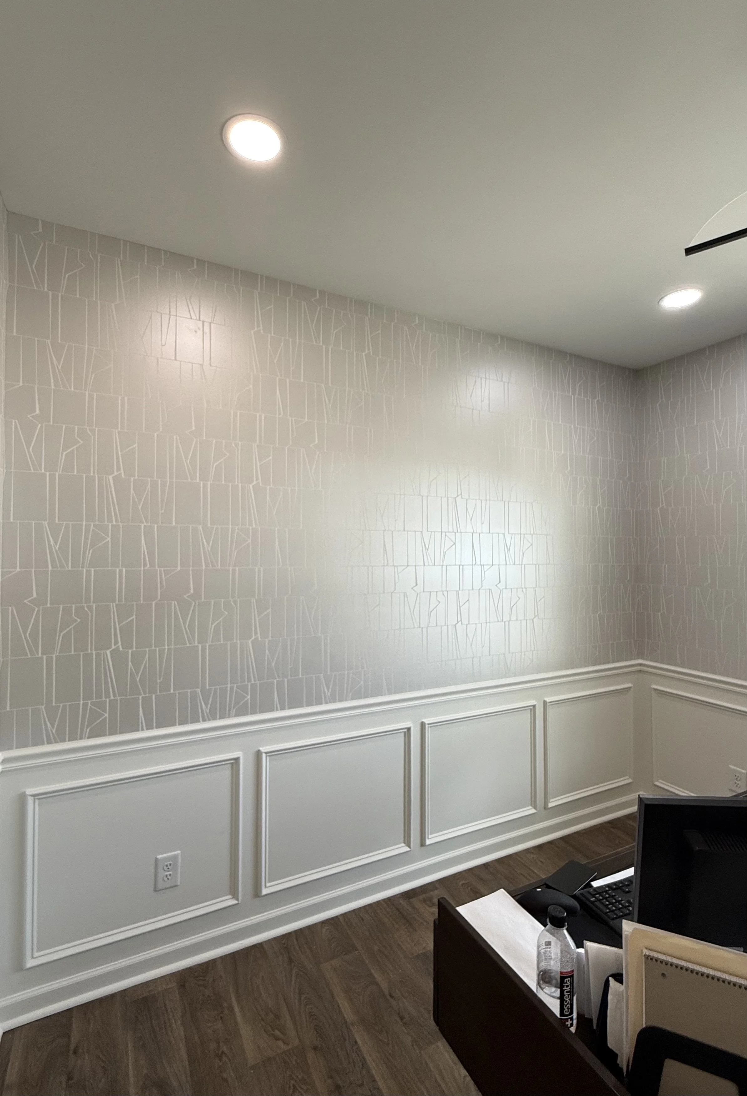 Office room with beige patterned wallpaper, white wainscoting, wood flooring, and ceiling lights. Part of a desk with office supplies is visible in the lower right corner.
