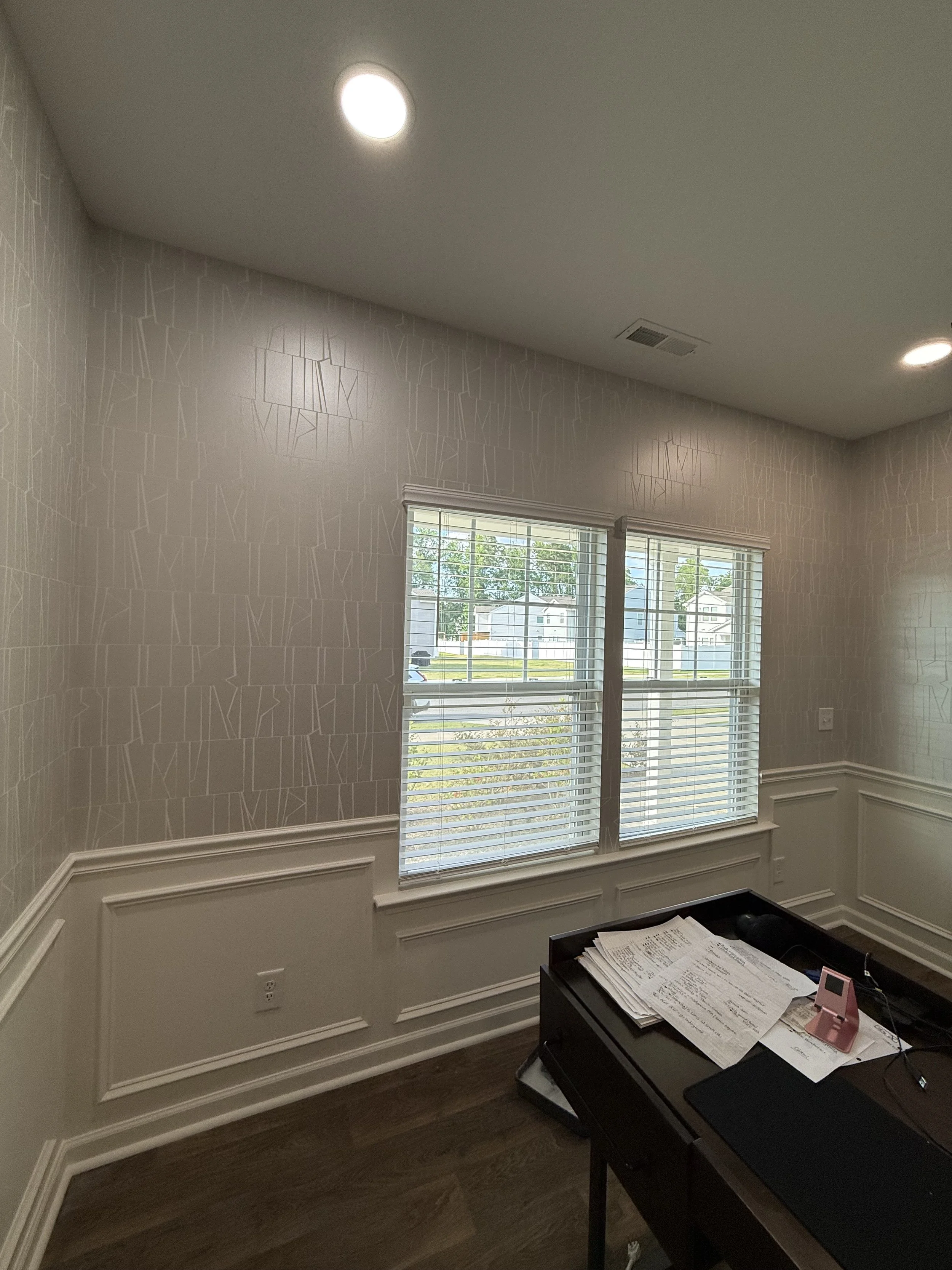 Interior of a room with two windows, beige patterned wallpaper, and white molding; a desk with papers and office supplies.