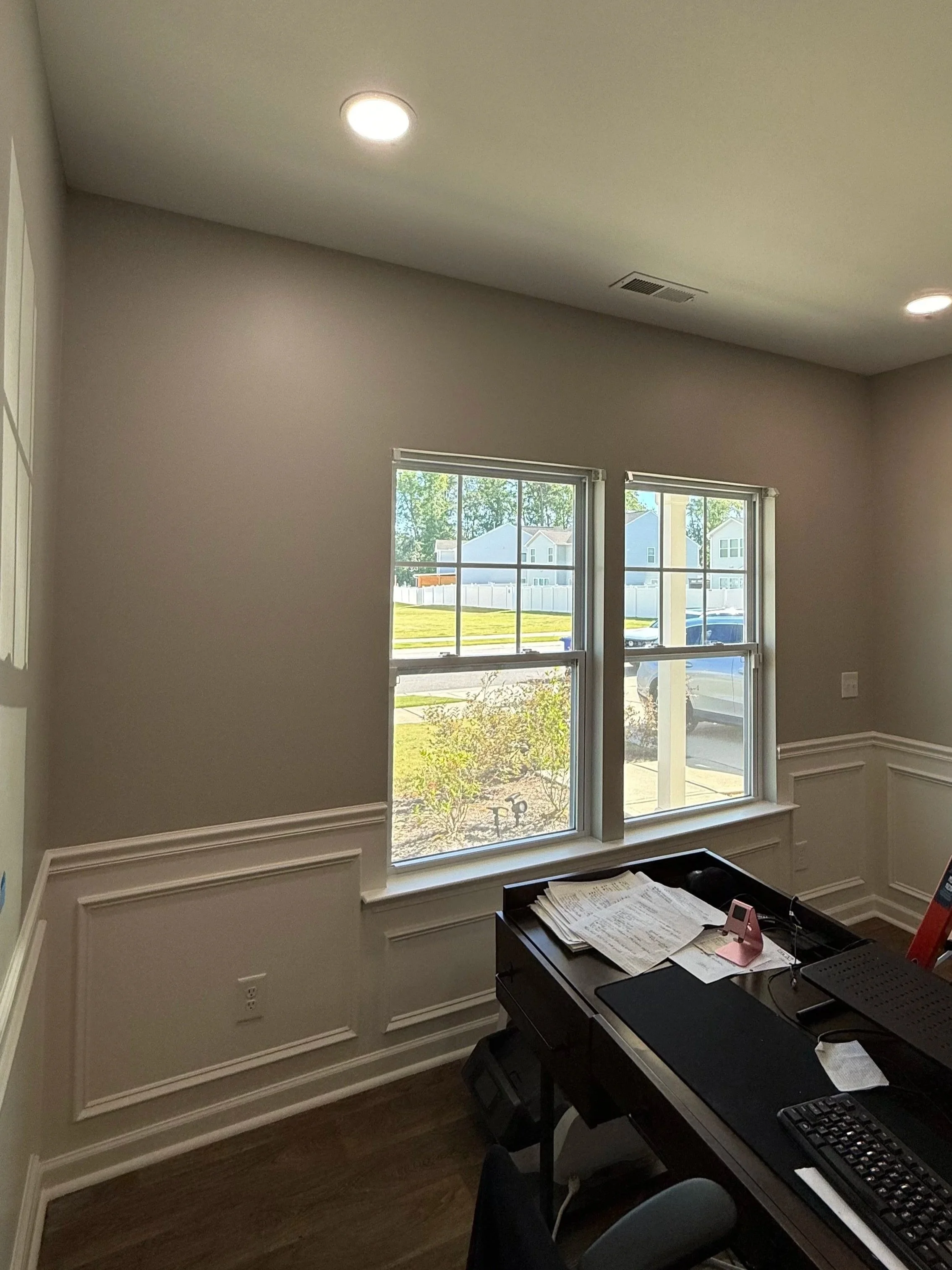 A room with a window looking outside to houses and trees, with a desk holding papers, a keyboard, a computer, and office supplies.