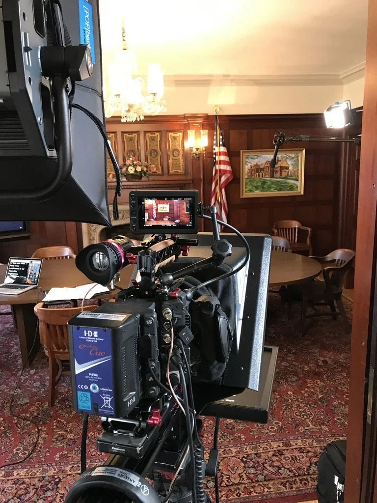 Recording setup in a room with a large round table, wooden chairs, an American flag, painting on the wall, and a chandelier.