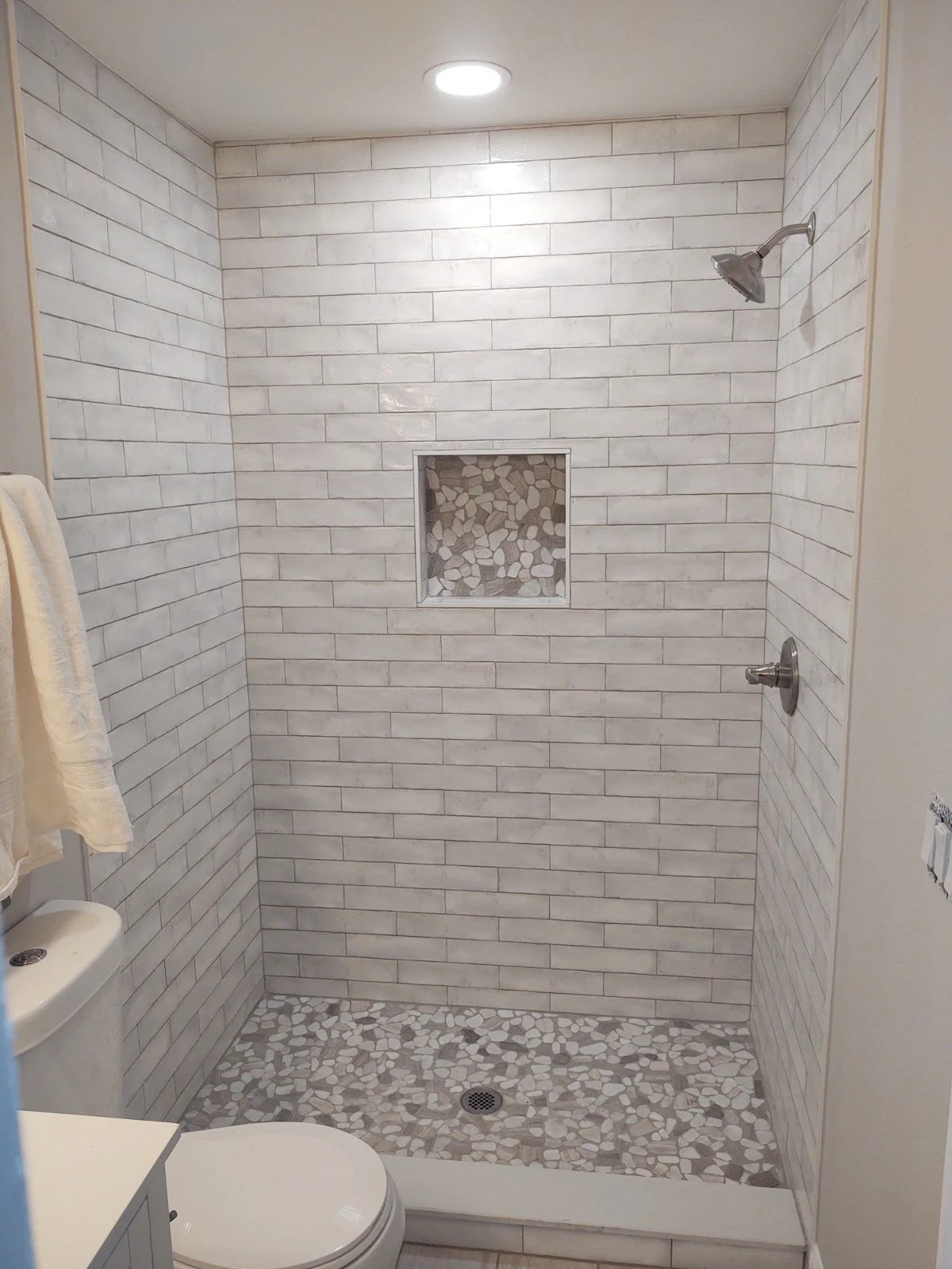A modern shower stall with white subway tile walls, a central alcove with patterned stones, a pebble tile floor, a wall-mounted showerhead, and a ceiling light.