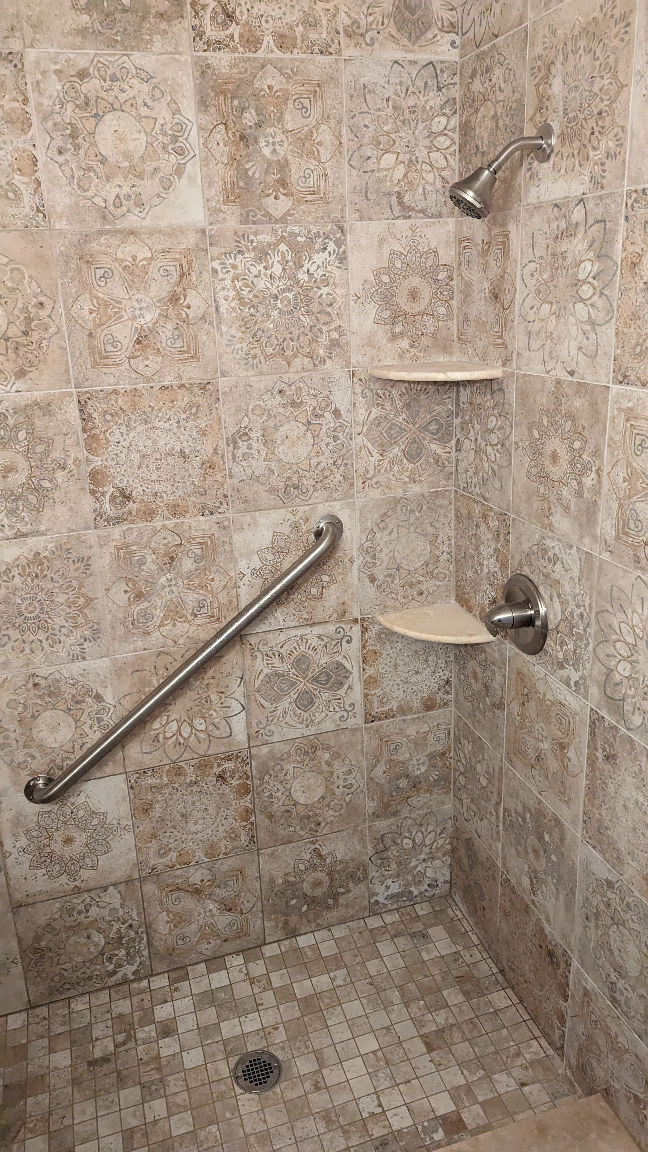 A shower stall with patterned tile walls, a metal grab bar, metal showerhead, and two small corner shelves.