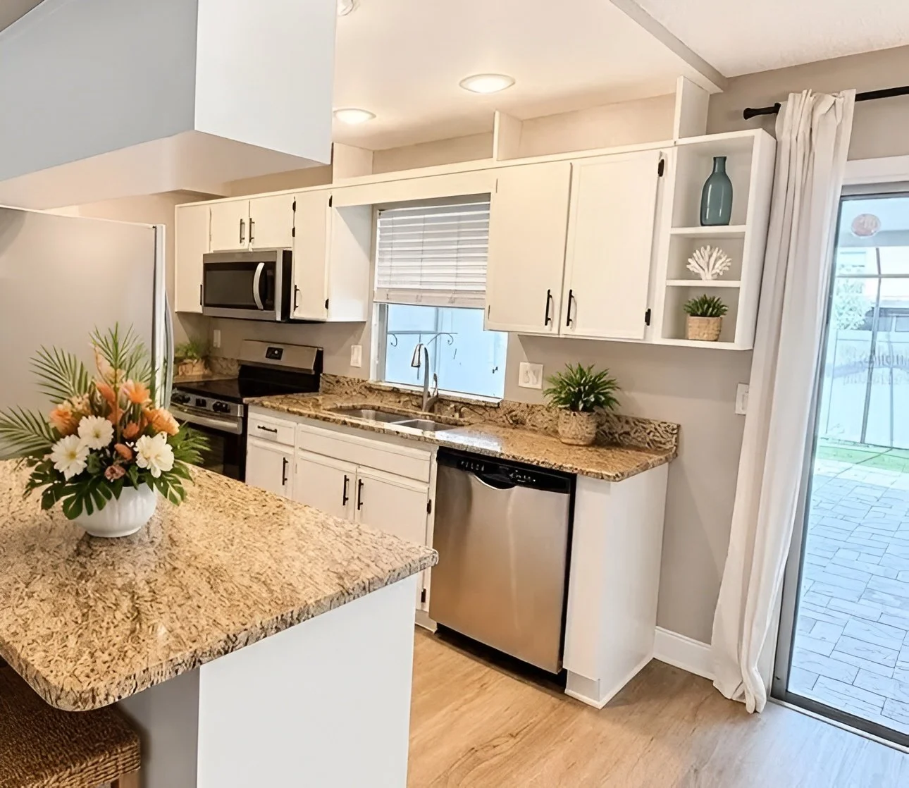 Kitchen with white cabinets, granite countertops, stainless steel appliances, a window with blinds, and sliding glass door with patio outside. Decor includes a flower arrangement and small potted plants.