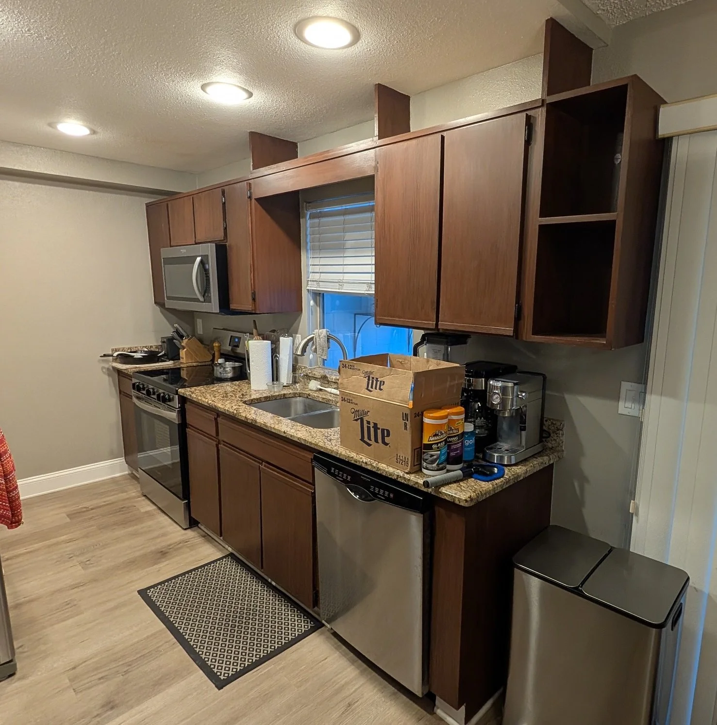 Kitchen with brown cabinets, granite countertops, stainless steel stove and microwave, small sink, and a cardboard box on countertop containing cleaning supplies and a coffee machine. There is a black and white rug on wood floor.