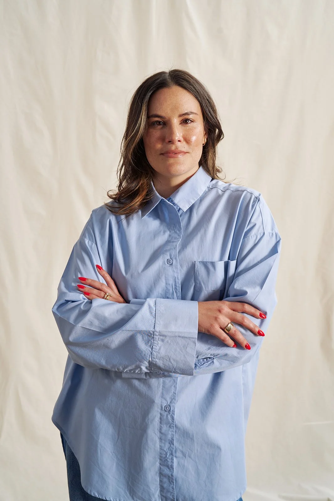 A woman with shoulder-length brown hair wearing a light blue button-up shirt with crossed arms, standing in front of a plain curtain background.