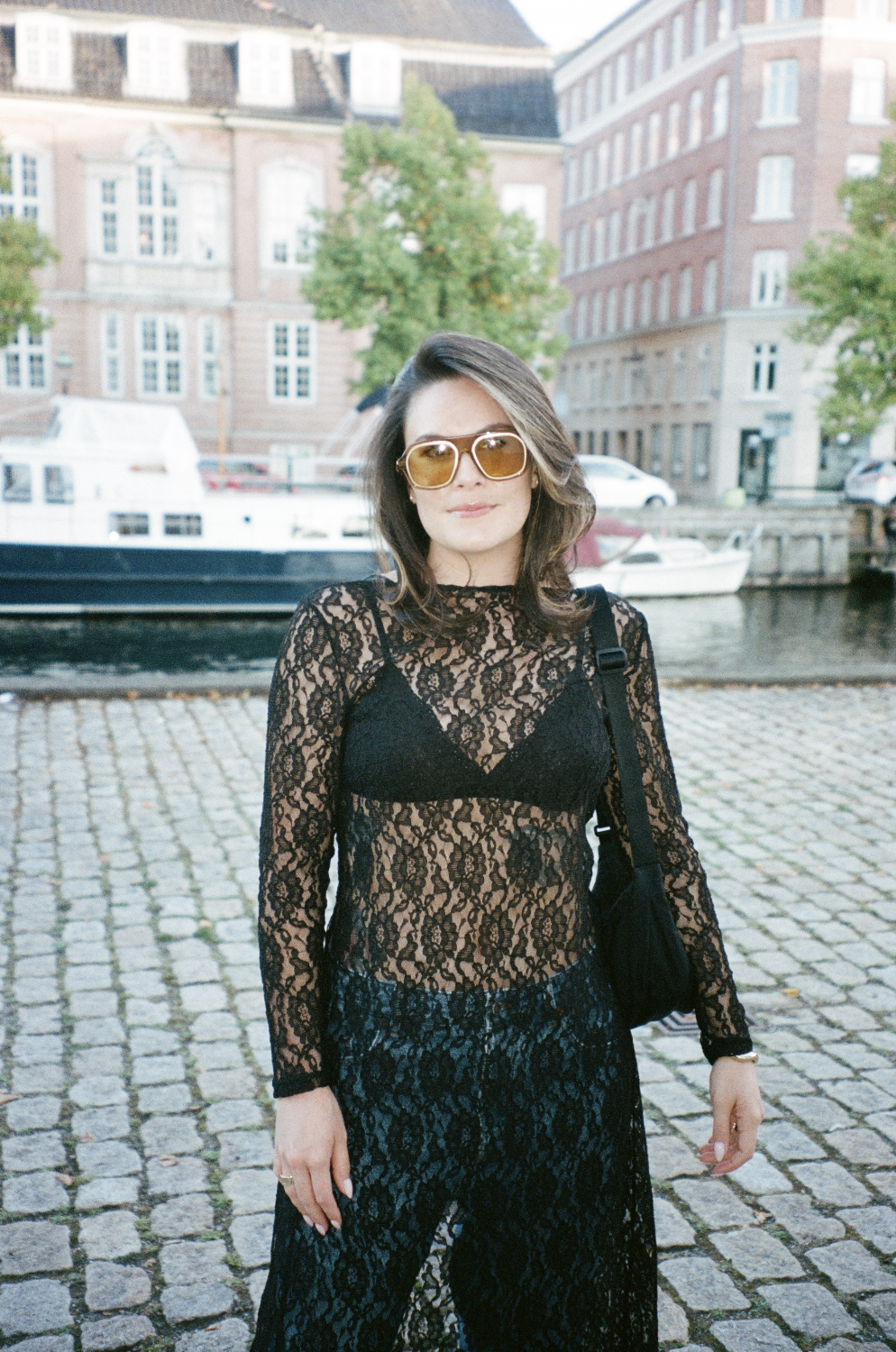 A woman wearing a black lace dress, tan sunglasses, and carrying a black bag, standing on cobblestone pavement near a canal with boats, trees, and historic brick buildings in the background.