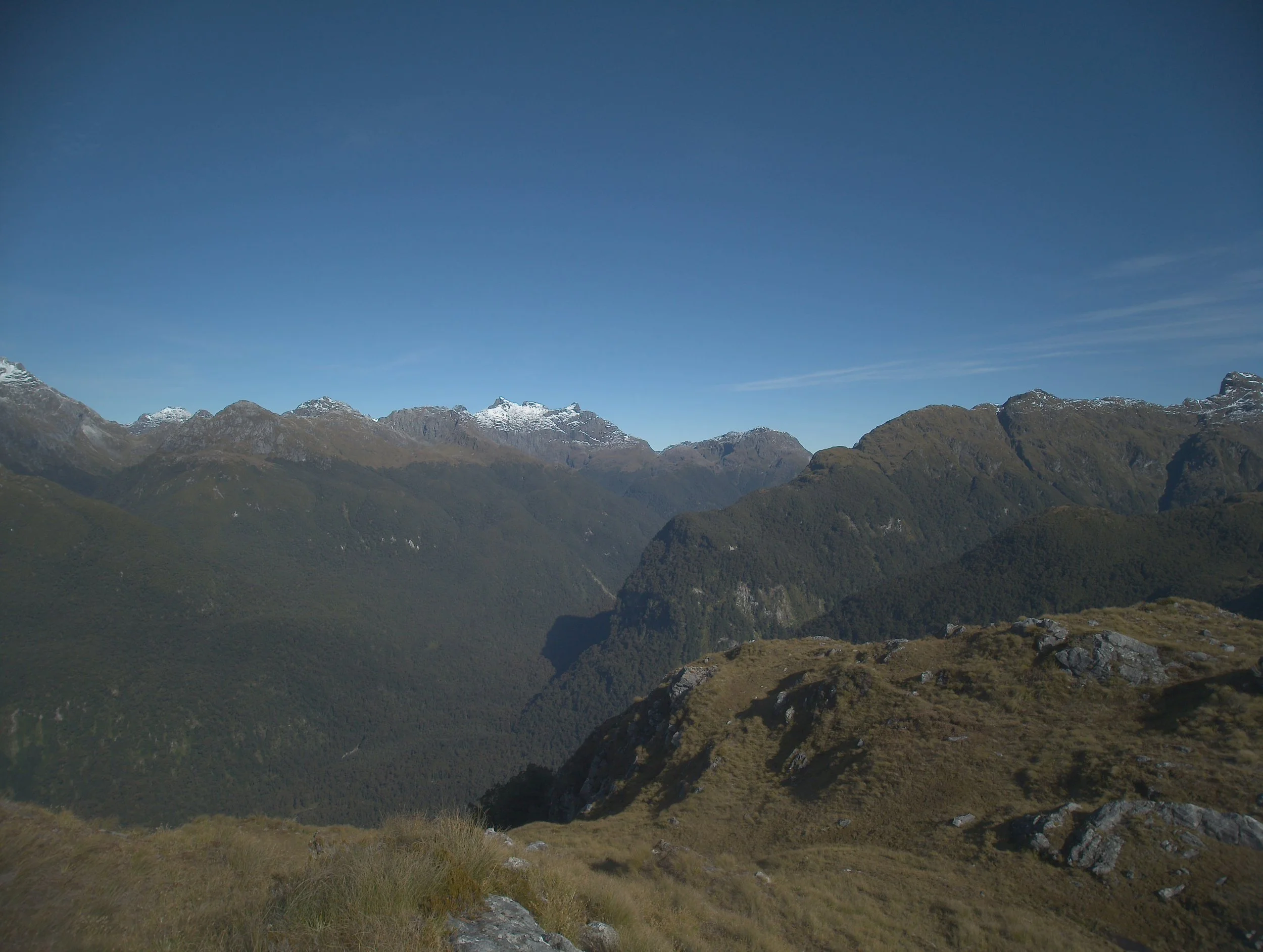 Mountain range with snow-capped peaks under a clear blue sky, green hills in the foreground.