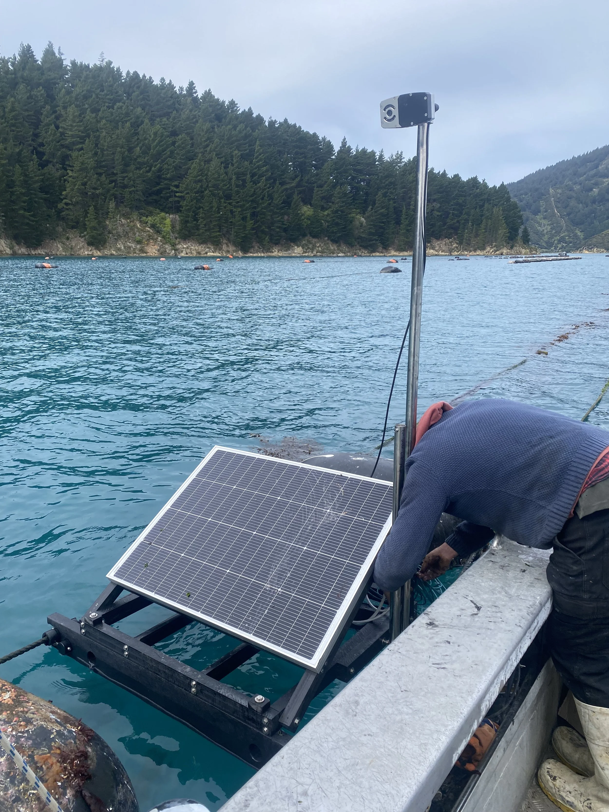 Person installing a solar panel on a boat dock with a scenic backdrop of water, trees, and hills.