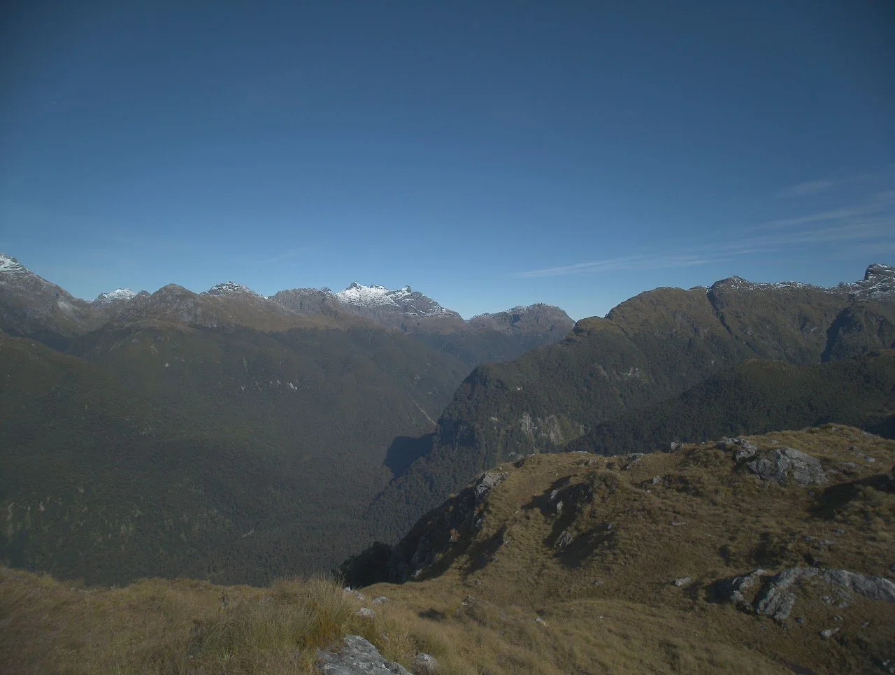 Scenic mountain landscape with green hills in the foreground, tall rugged mountains with some snow-capped peaks in the background, and a clear blue sky.