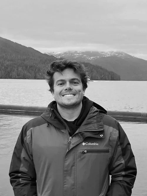 A young man smiling outdoors, wearing a Columbia jacket, standing in front of a lake with mountains in the background.