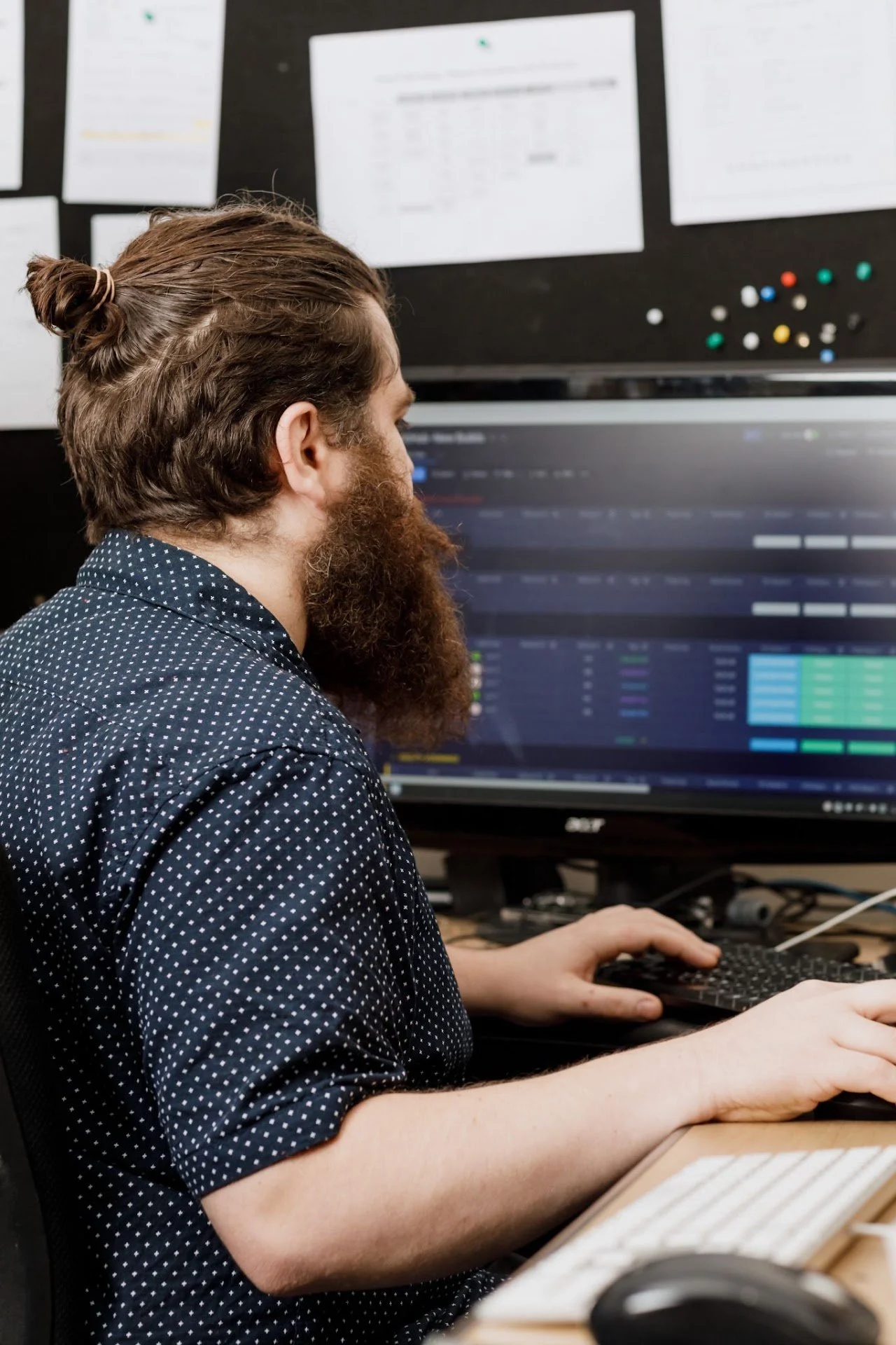 A man with long hair tied in a bun and a full beard is sitting at a desk working on a computer, with multiple sheets of paper pinned to a black backdrop on the wall behind him.