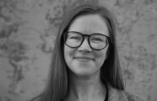 A woman with long hair and glasses smiling in front of a textured background.