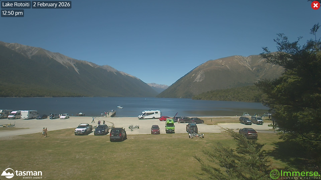 A lakeside scene at Lake Rotoiti on February 2, 2026, at 12:50 pm, showing a parking area with various cars, a camper van, and people near the water. The lake is surrounded by green mountains under a clear blue sky.