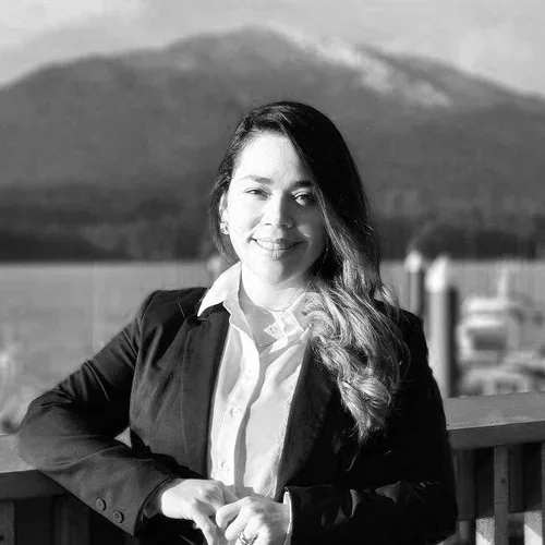 A young woman with long hair standing on a balcony with a mountain and parking lot in the background, smiling in black and white.