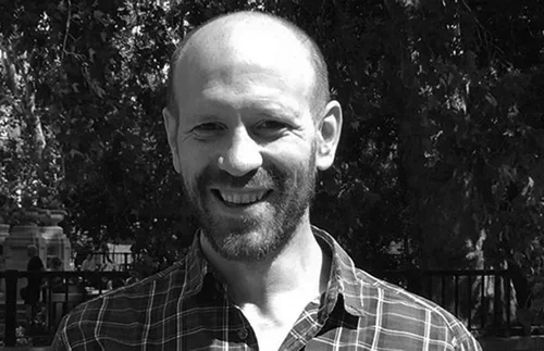 A smiling man with a beard and short hair, wearing a checkered shirt, outdoors in front of trees.