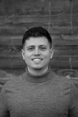 A young man smiling, wearing a turtleneck sweater, standing in front of a wooden wall.