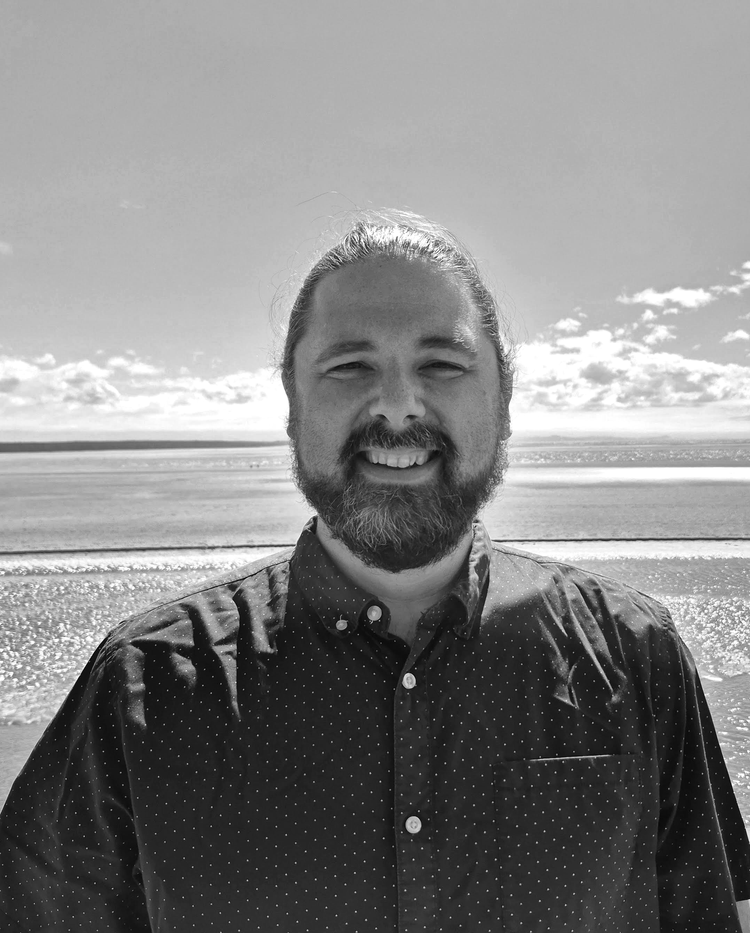 A man with a beard and long hair smiling on a beach with the ocean and sky in the background.