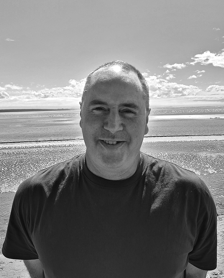 Black and white photo of a smiling man standing on a beach with the ocean and partly cloudy sky in the background.
