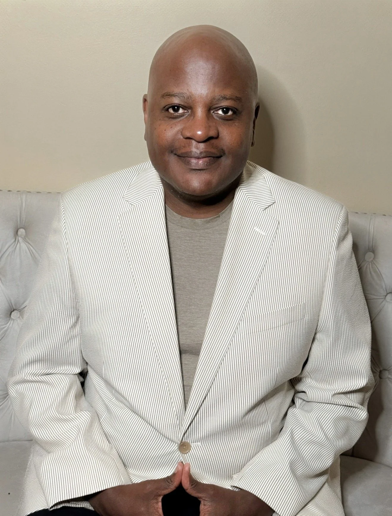 Portrait of a smiling Black man with a shaved head, wearing a light-colored striped blazer over a gray shirt, sitting on a white tufted couch against a plain beige wall.