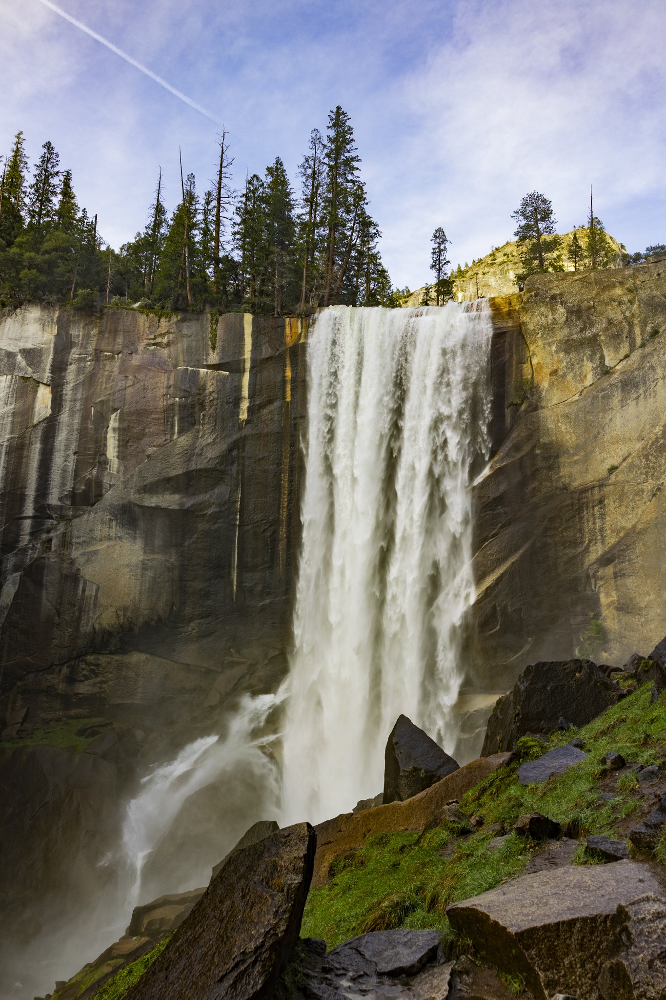 Waterfall cascading down a rocky cliff with trees at the top and rocks at the base, with a blue sky and wispy clouds above.