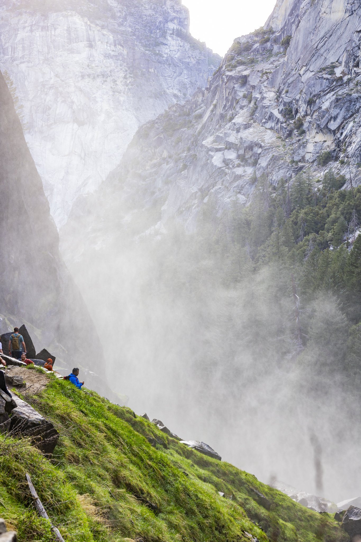 People sitting on a grassy slope near a waterfall in a canyon with cliffs covered in trees.