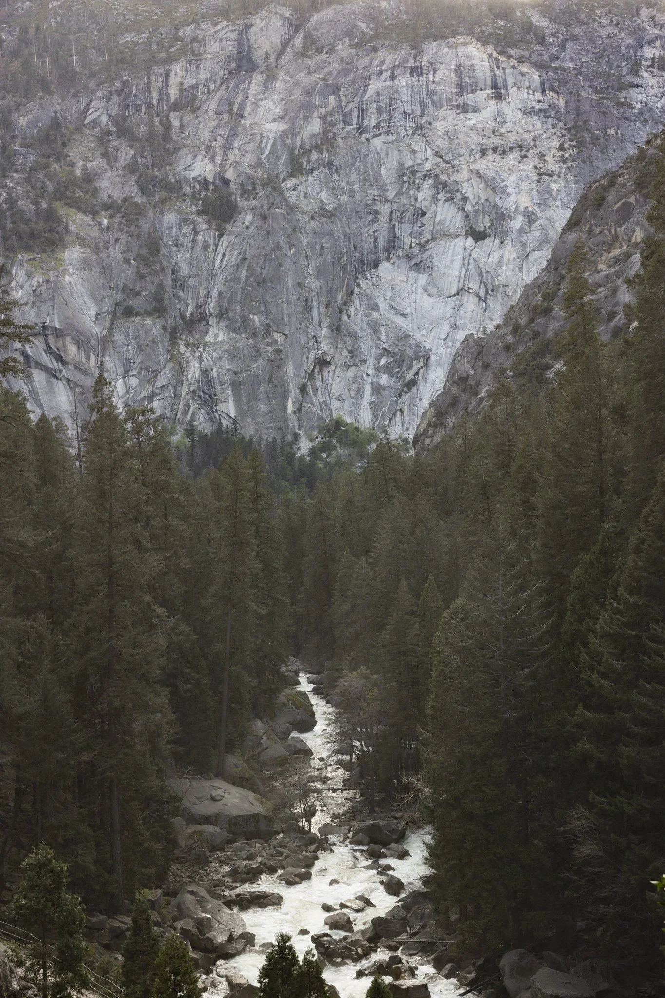 A mountainous landscape with steep gray rock faces and a flowing river at the bottom, surrounded by dense green trees.