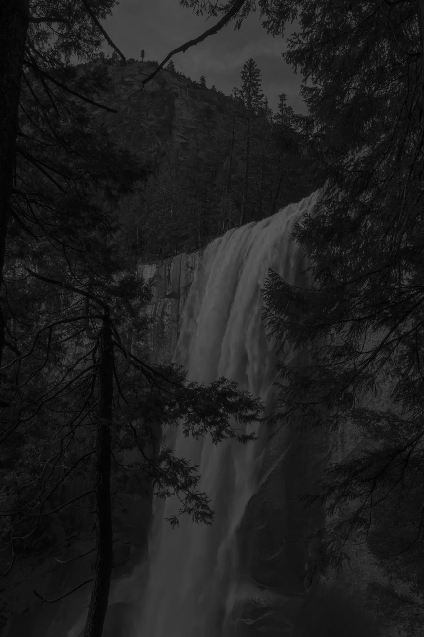 A dark, black-and-white photo of a tall waterfall surrounded by trees, with a rocky hill and trees in the background under a cloudy sky.