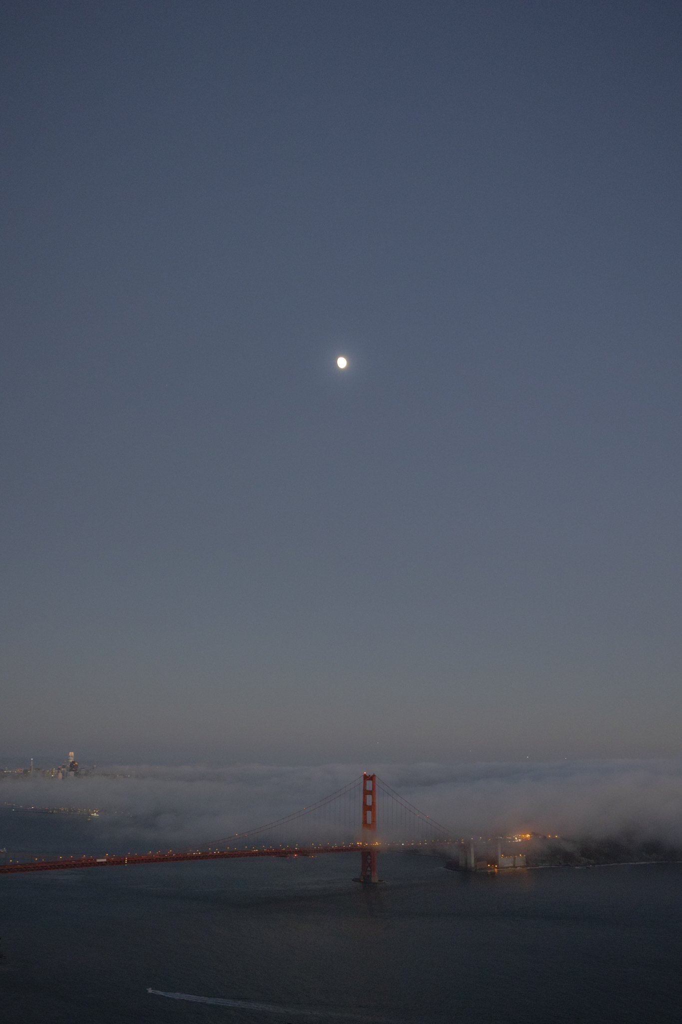 Night view of the San Francisco-Oakland Bay Bridge with the moon in the sky and fog over the water.