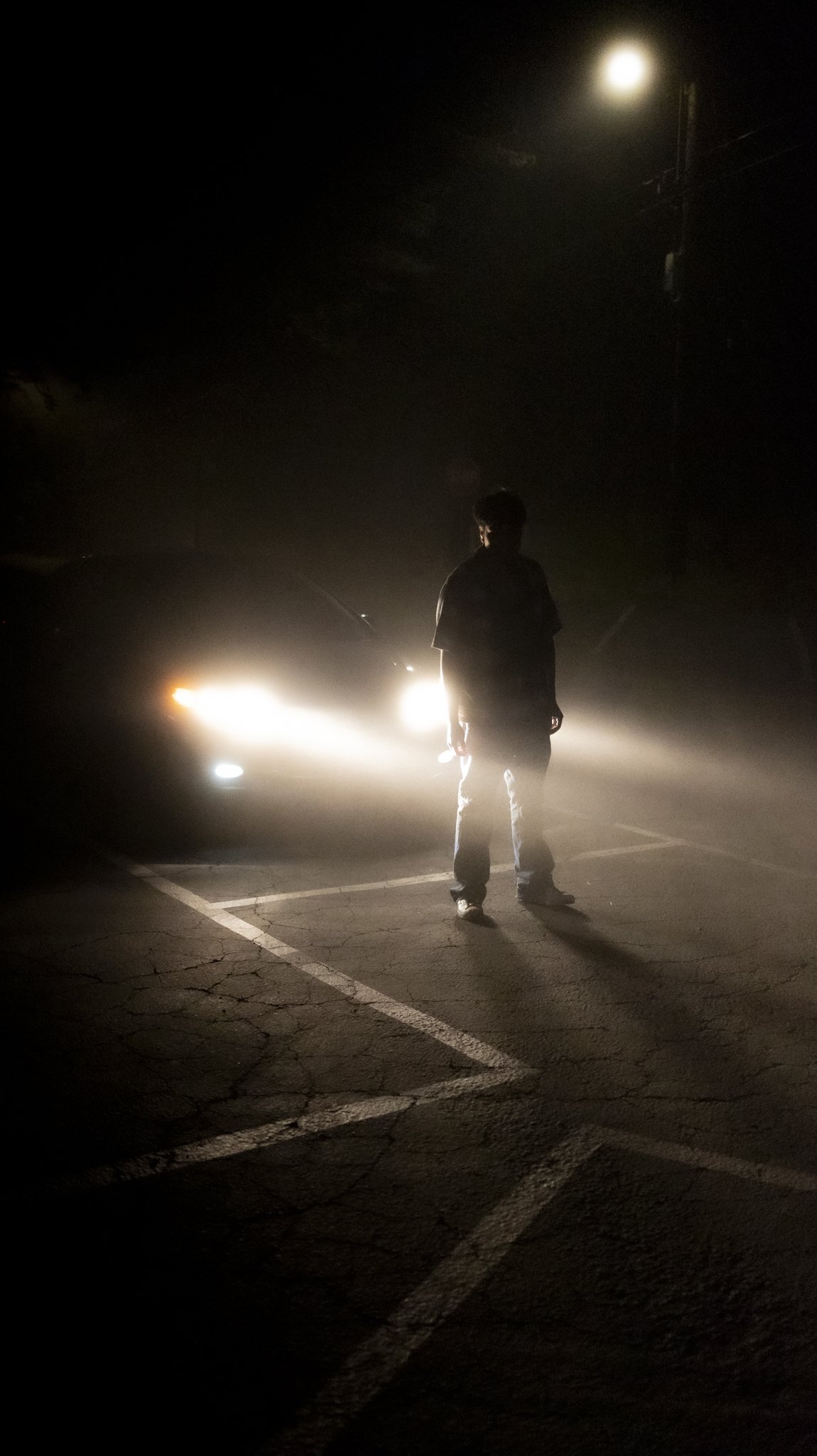 Silhouette of a man standing in a parking lot at night, illuminated by headlights of a car and a bright moon in the sky.