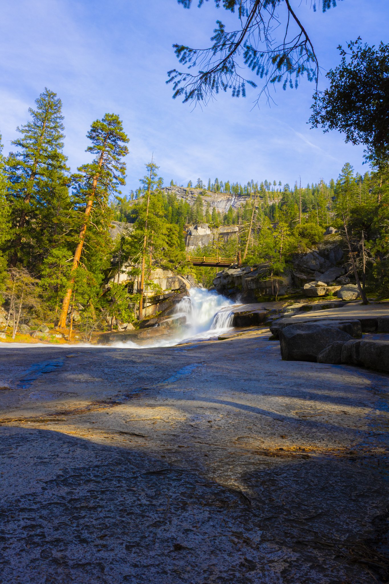 A scenic outdoor view of a waterfall flowing over rocks in a forest, with tall pines and a blue sky in the background.