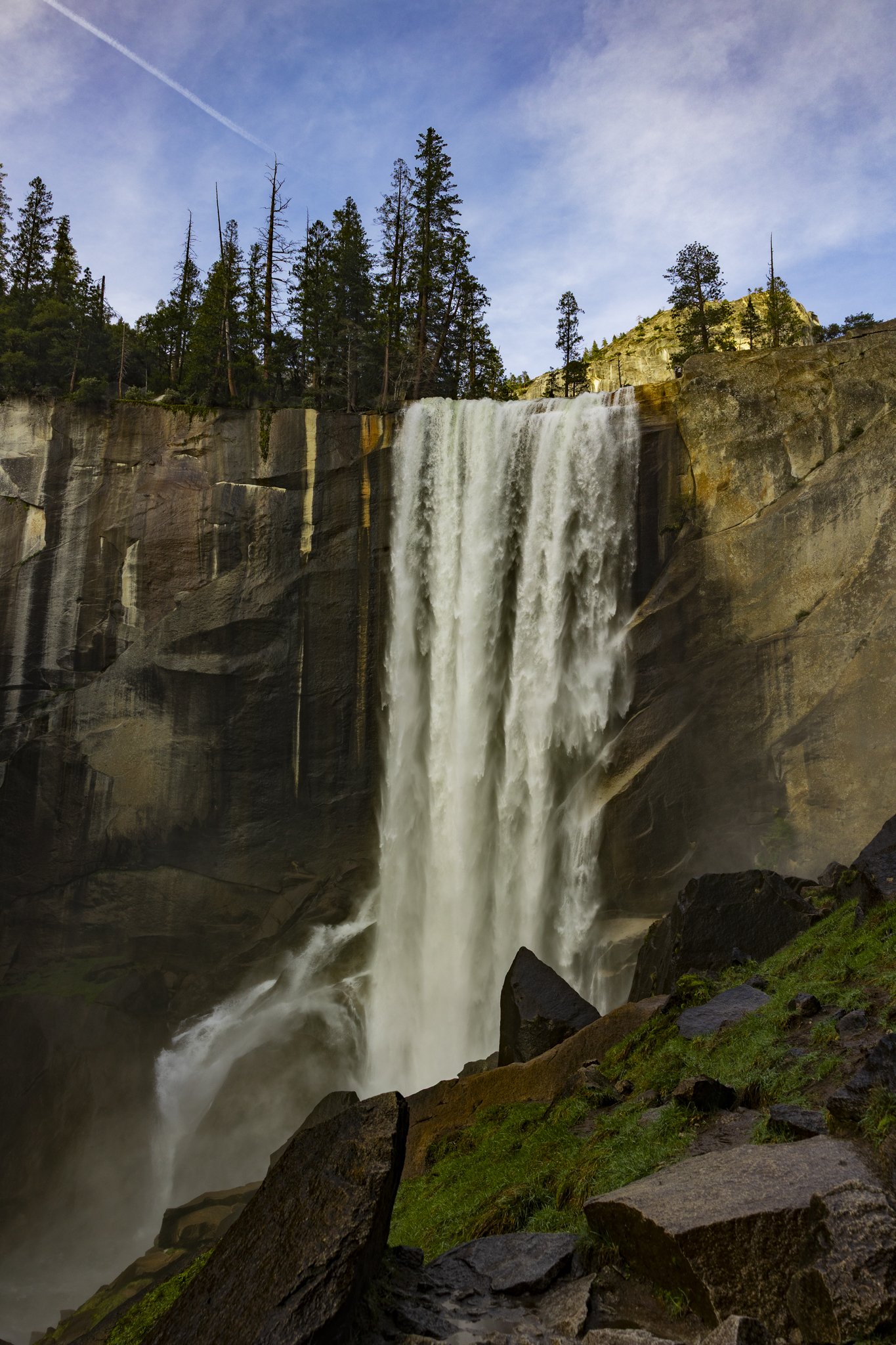 A scenic view of a waterfall cascading down a rocky cliff with pine trees on top and a blue sky with a few clouds in the background.