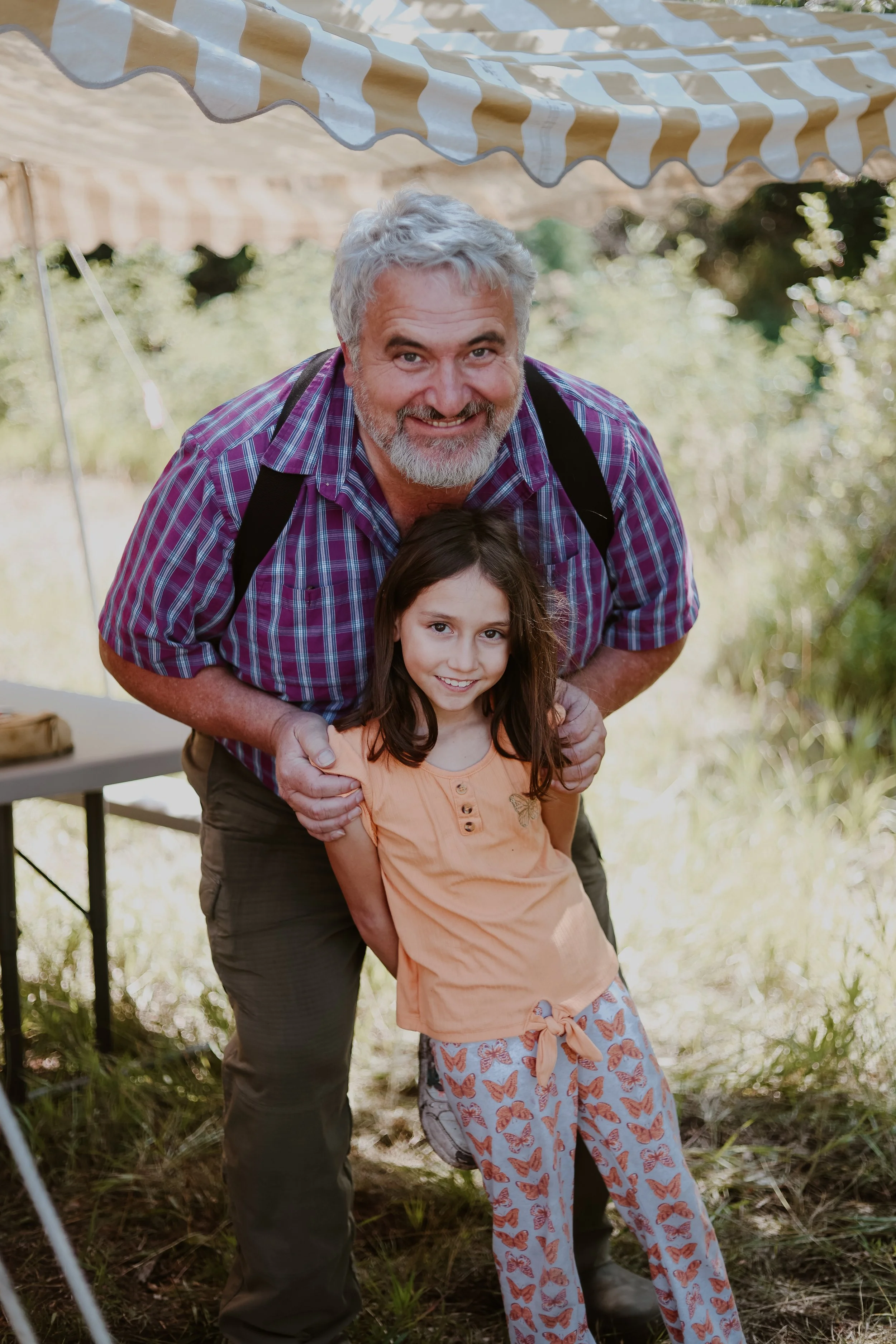 An older man with gray hair and a beard, wearing a plaid shirt, and a young girl with long brown hair, wearing a peach-colored shirt and butterfly-patterned pajama pants, posing outdoors under a striped yellow and white canopy.