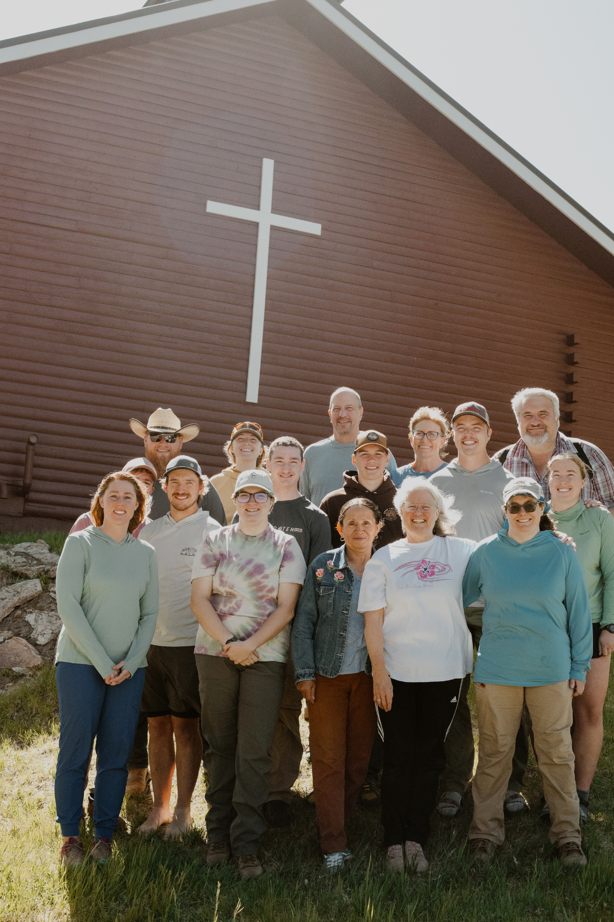 Group of people smiling and standing in front of a brown church with a large white cross on the wall, outdoors on a sunny day.