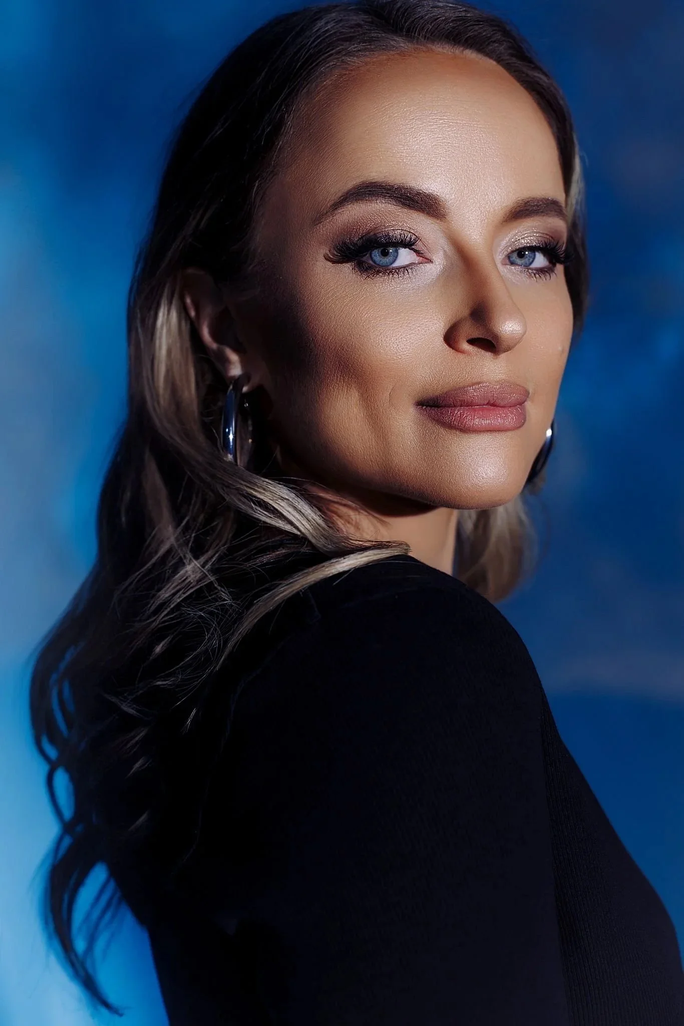 Close-up portrait of a woman with long, wavy hair, blue eyes, and subtle makeup, dressed in a black top against a blue background.