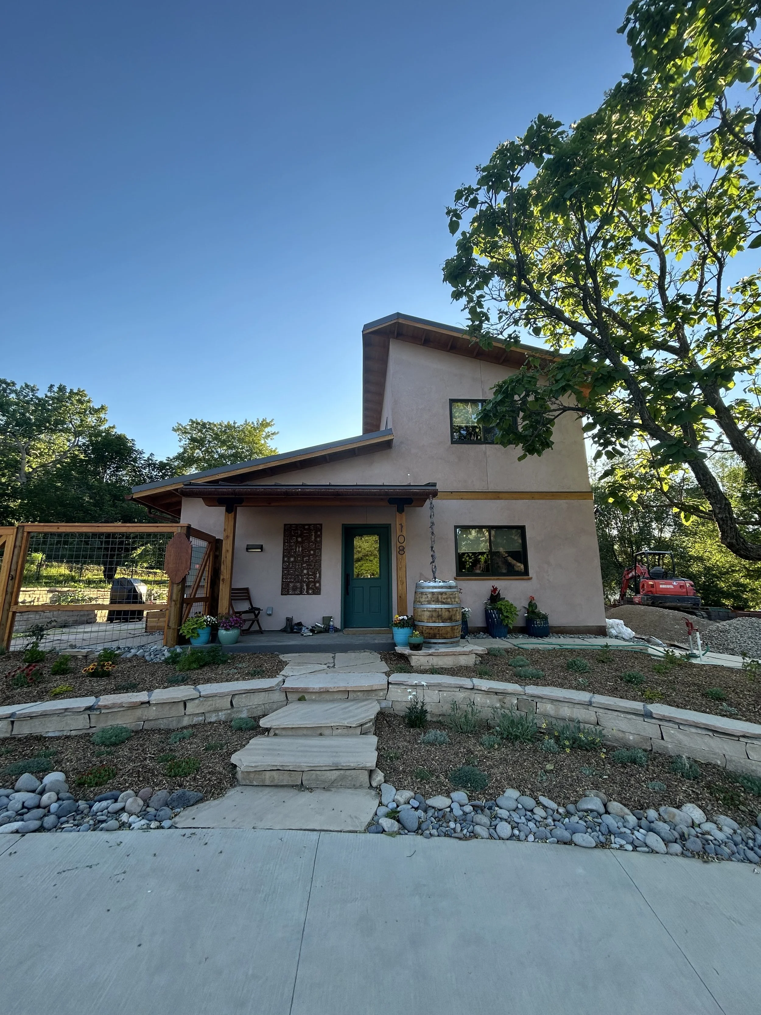 Front view of a modern two-story house with a garden in front, stone steps leading to the door, and a red compact utility vehicle parked on the right side. The house has a beige exterior with black window frames and a teal door, surrounded by trees a