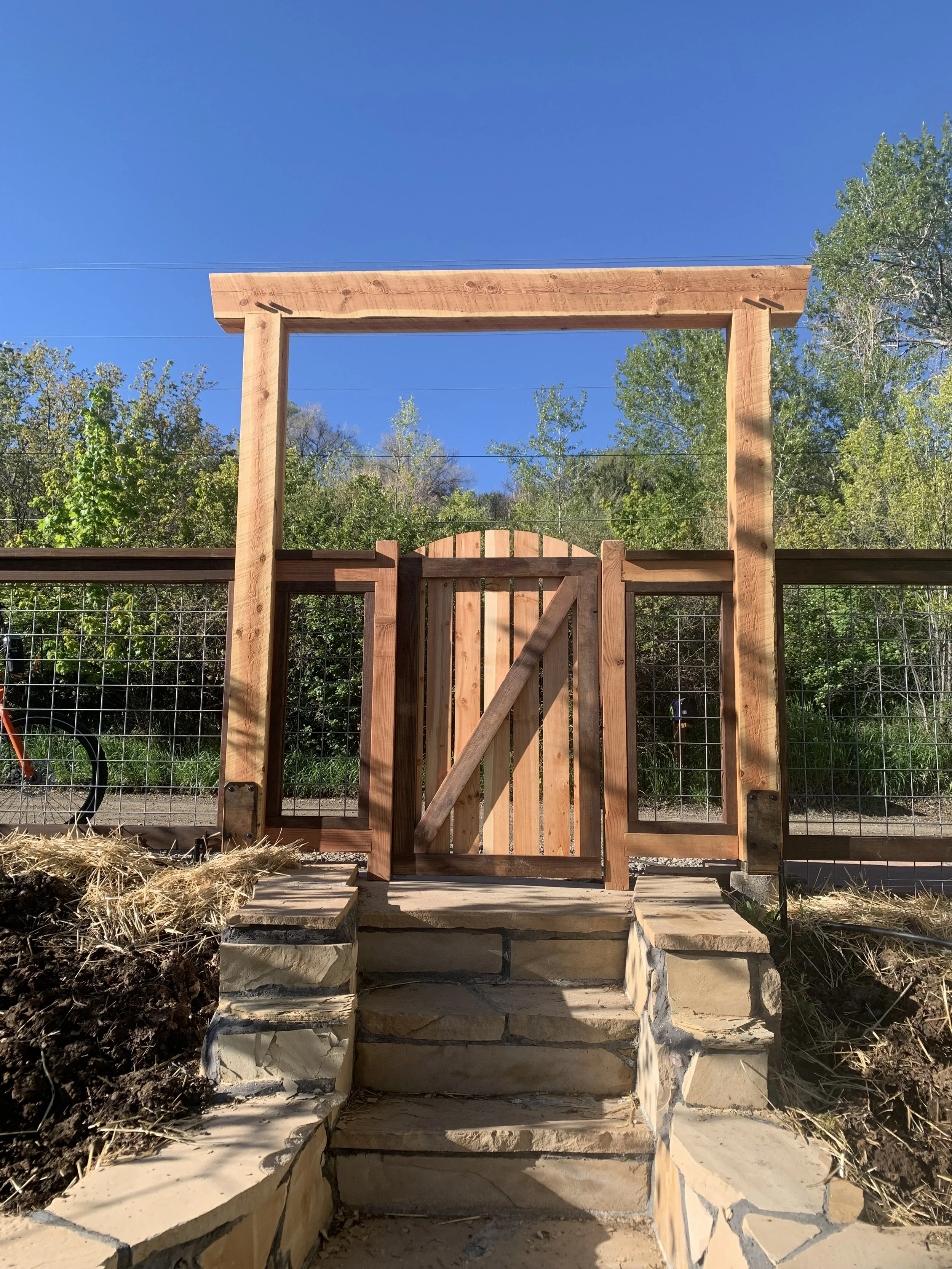 Wooden gate under construction, with steps leading up to it, surrounded by a wire fence, with trees and blue sky in the background.