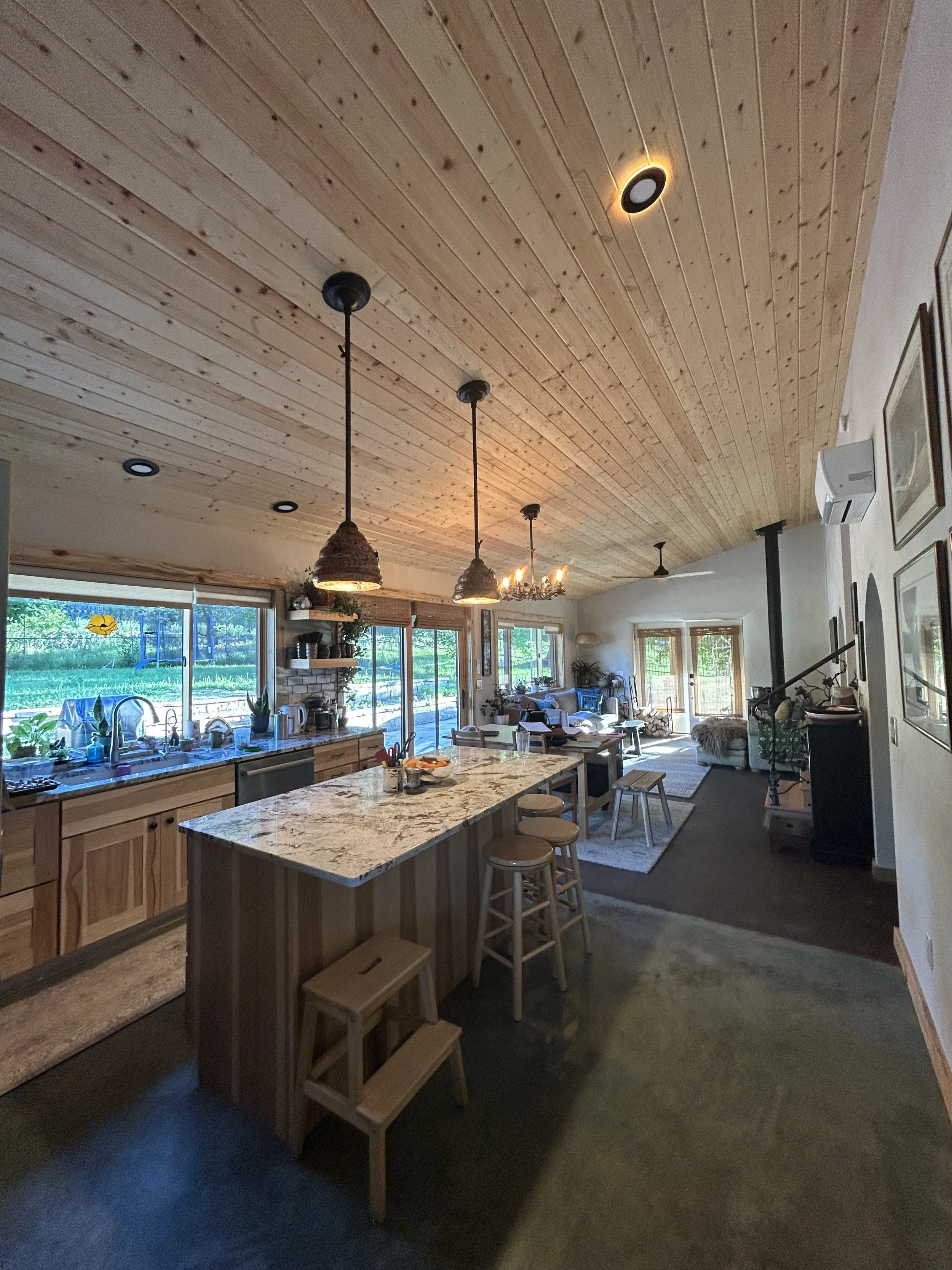 Open-concept living area with a kitchen and dining space, featuring a wooden ceiling, pendant lights, a fireplace, large windows, and various furniture and decorations.